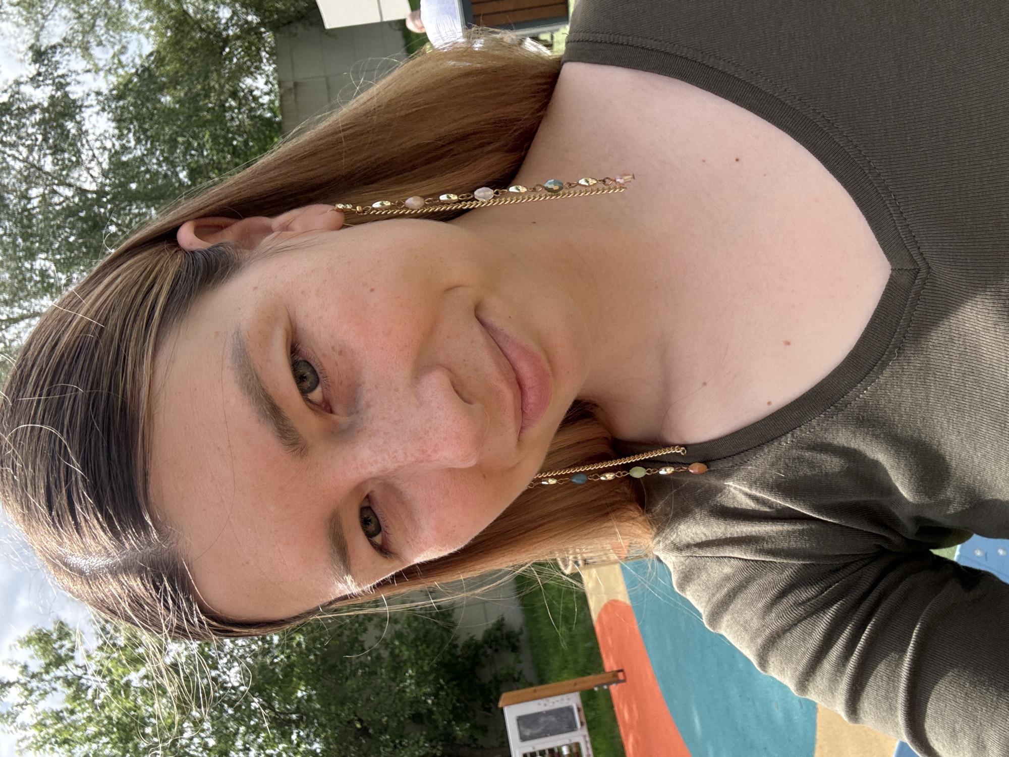 A woman is facing the camera with a slightly tilted head. She has fair skin, light brown hair, and green eyes. She is wearing a dark green V-neck shirt and long earrings with beads. The background includes trees, a building, and a play area with blue and orange surfaces. The location is unspecified.
