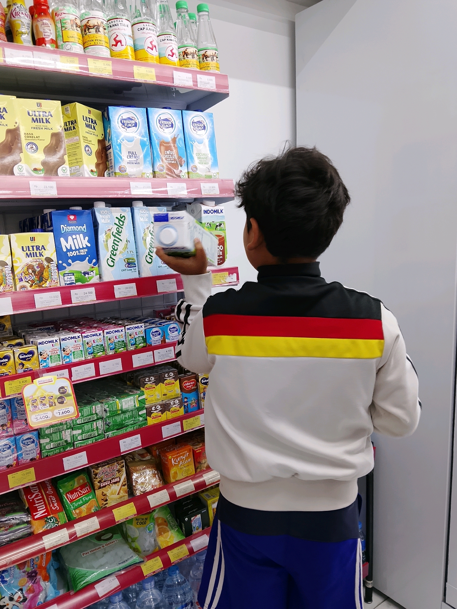 A young boy, viewed from the back, stands in an aisle of a brightly lit convenience store, likely Alfamart given the branding on the shelves. He is wearing a white jacket with black, red, and yellow stripes across the chest, and blue shorts. His right hand is extended, holding a white and blue carton of milk, possibly Indomilk or Diamond Milk. He is in the process of selecting or examining the product. The shelves are stocked with a variety of beverages and food items, including milk cartons of different brands such as Ultra Milk, Frisian Flag, and Greenfields, as well as bottles of what appear to be Indonesian traditional drinks like larutan penyegar. The price tags visible are in Indonesian Rupiah (Rp). The overall environment suggests a typical retail setting in Indonesia.