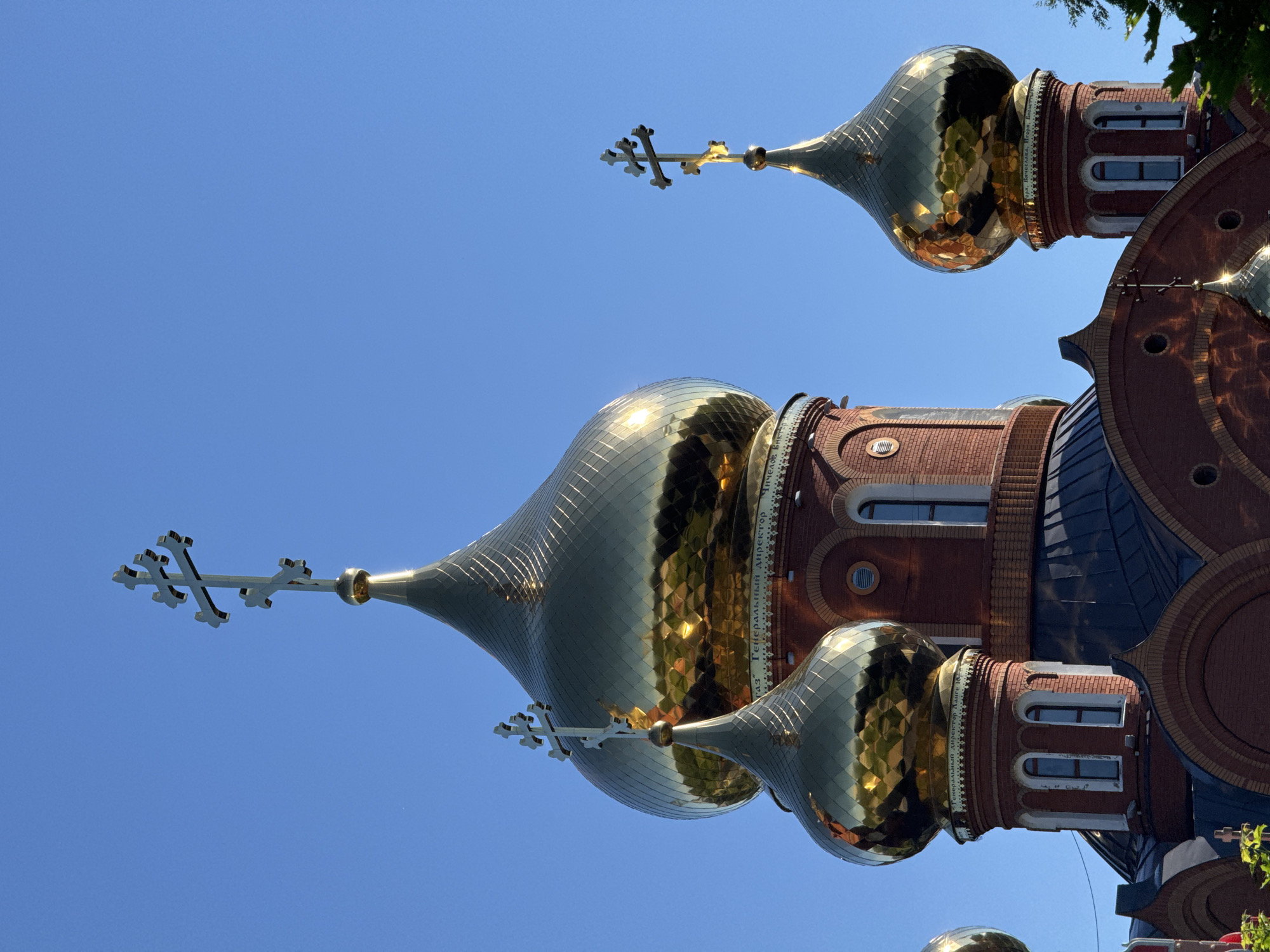 The image shows a close-up of golden domes and crosses of an Orthodox church against a clear blue sky. The domes are bulbous and covered in reflective, faceted gold plating, creating bright highlights and reflections. Several brick towers support the domes, featuring arched windows with white trim. The brickwork is a deep red-brown. Inscriptions in Russian lettering are visible on a band around one of the dome bases. The lighting suggests it is daytime, likely morning or afternoon, with the sun illuminating the domes from the side. The church appears to be the Church of the Intercession of the Holy Virgin in Chaikovsky, Russia, given the distinctive architecture and context of Chaikovsky being known for its Orthodox churches. No people are visible in the image.