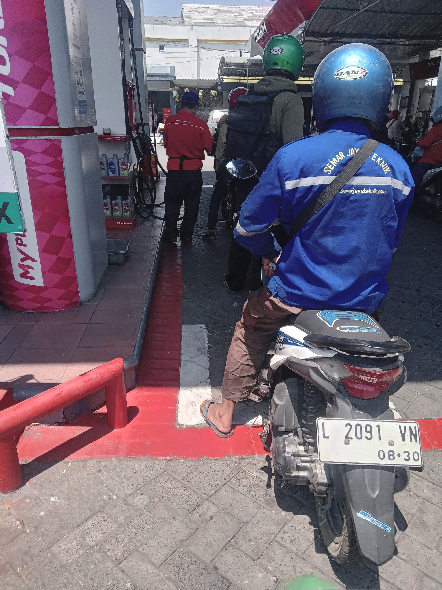A medium shot captures a street scene at a gas station in Sawotratap, Indonesia, under bright daylight. In the foreground, a person wearing brown pants and a blue jacket with white stripes is seated on a white and grey motorcycle, facing away from the camera. The blue jacket has text on the back that reads "SEMAR JAYATEKNIK" and a website address "_semarjayateknik.com". A black strap is slung across the person's right shoulder. The motorcycle has a white license plate displaying "L 2091 VN" and "08.30". A sticker with blue lettering on a white background is visible on the rear fender. The person is wearing grey flip-flops.
The motorcycle is positioned on a grey paved area with red and white painted lines indicating a pathway or boundary. To the left of the motorcycle, a red metal railing is visible. Behind the railing, a large pink and white structure, possibly part of the gas station's display, is present with some text that is partially obscured.
Further in the background, several other individuals are present, some on motorcycles. One person in a red uniform stands near a gas pump. Another individual wears a green helmet, and someone else has a red helmet. Gas pumps and awnings are visible, suggesting a commercial area. The environment appears to be a typical Indonesian gas station on a sunny day.