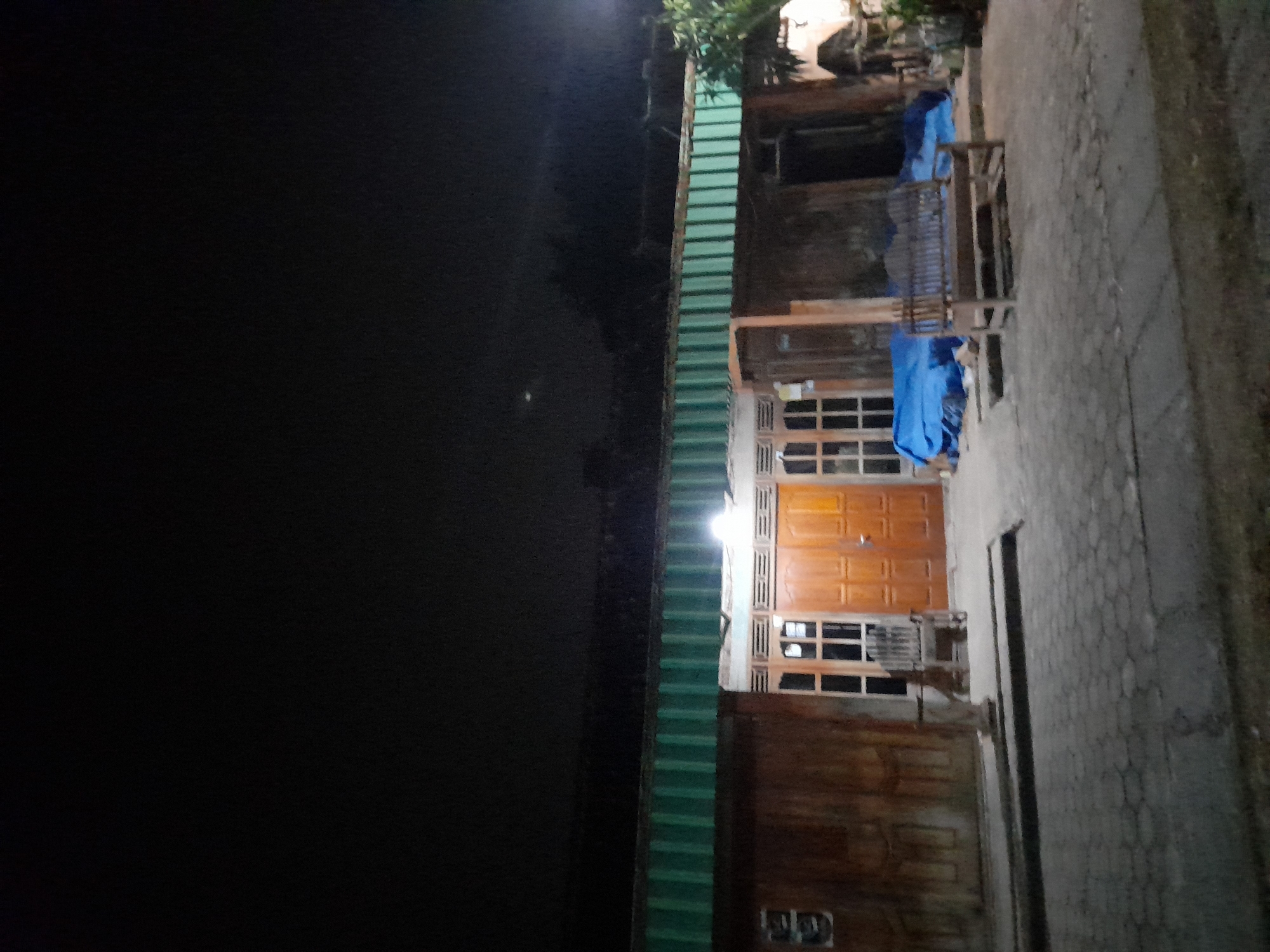 The image depicts a nighttime scene outside a building in Pulokulon, Indonesia. The building appears to be a house or a shop, with wooden walls and a green corrugated metal roof. In front of the building, there are several elements visible. A wooden door with glass panes is centrally located, flanked by windows with a grid pattern. To the left of the entrance, a wooden chair is placed. A wooden bench sits on the right side of the image, in front of a blue tarp covering something. The ground is paved with interlocking concrete blocks, and a small patch of grass is visible in the foreground. The sky above is dark, indicating it is nighttime. There are no people visible in the image.