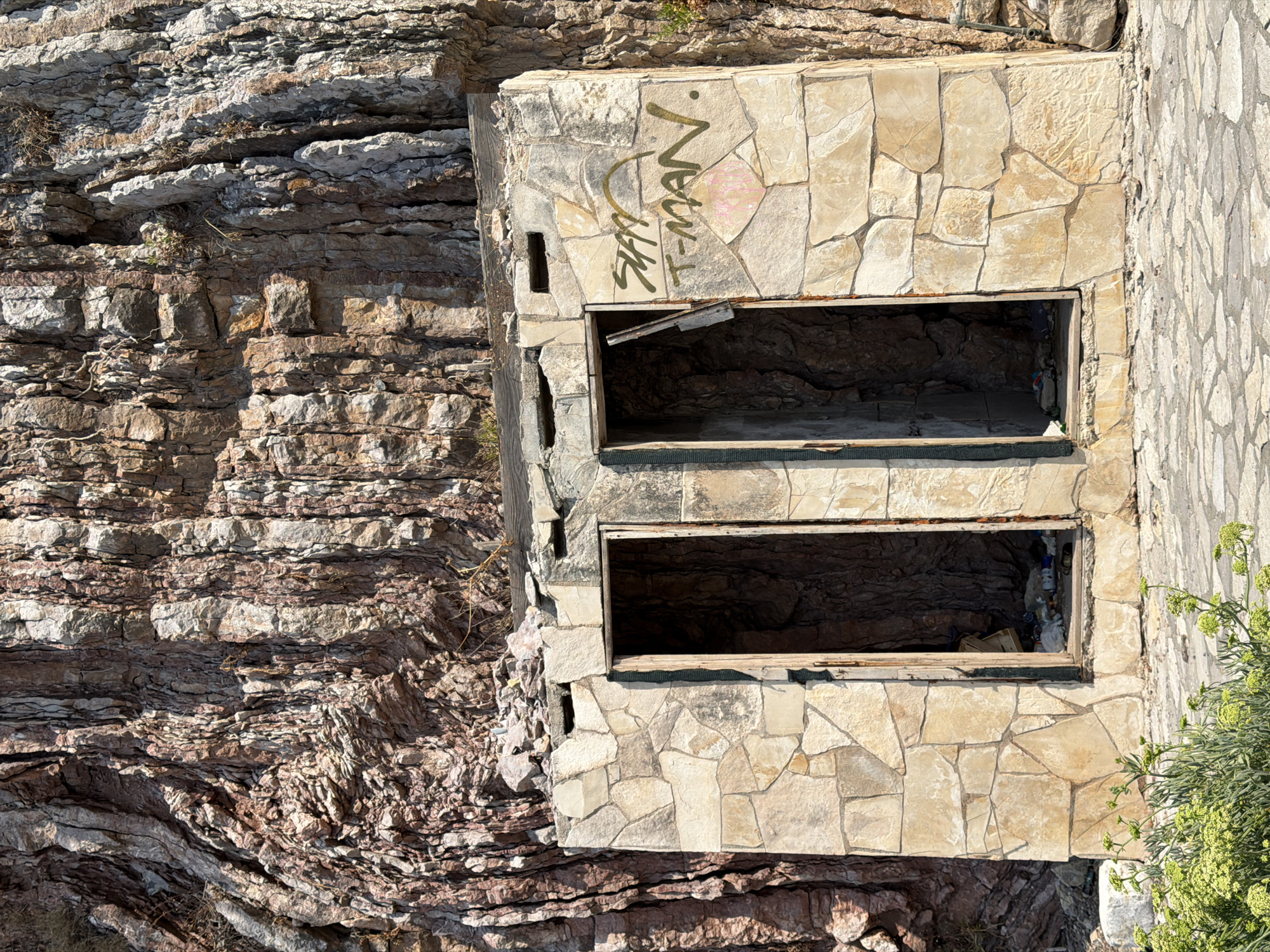 An abandoned stone structure with two openings stands against a large rock face in Boreti, Montenegro. The structure is built with irregularly shaped light-colored stones. The openings appear to be entrances to small rooms, with debris visible inside. One of the openings has a broken wooden door leaning against the frame. The stone facade on the right has graffiti in dark green spray paint that reads "STAY T-MAN." There are also hints of pink spray paint below the graffiti. The rock face behind the structure is reddish-brown and textured, with visible layers and crevices. The ground in front of the structure is paved with flat, irregularly shaped stones. Green foliage is visible in the lower left corner of the image. The scene suggests a derelict public facility in a natural, rocky environment.