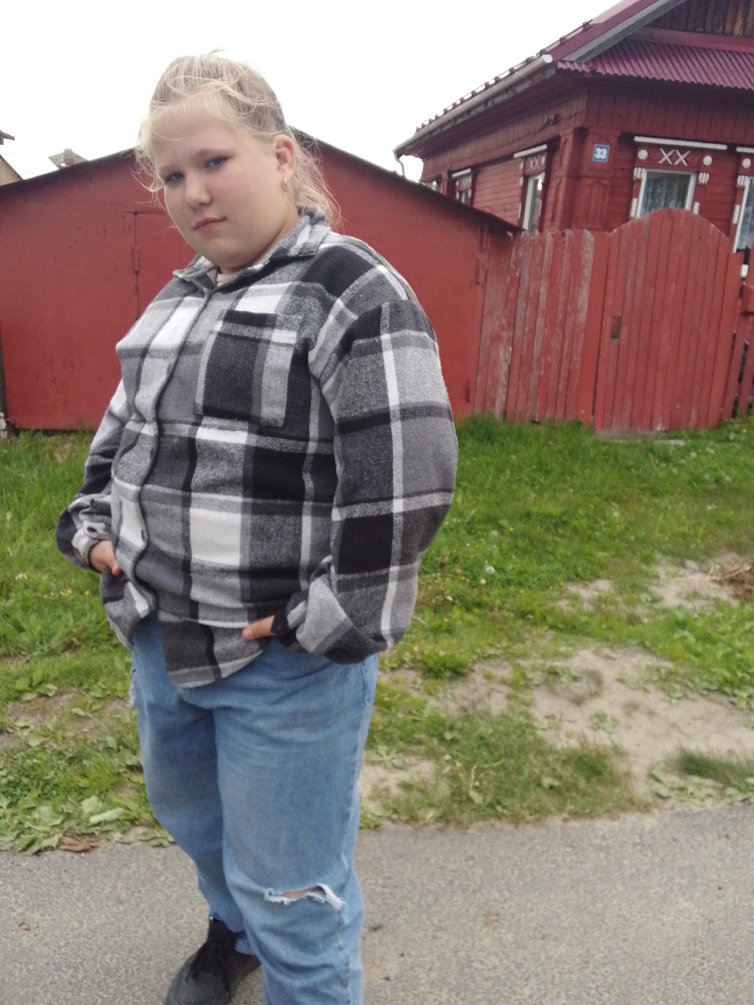 A girl stands in front of a red house and fence. The girl has blonde hair in a high bun and is wearing a gray and black plaid shirt with blue jeans that are ripped at the knee. She has both hands in her pockets. The house has a red roof and white trim. The fence is also red and has green grass on either side of it. There is a gray sidewalk or driveway in the foreground. The city location is Unknown, Unknown.