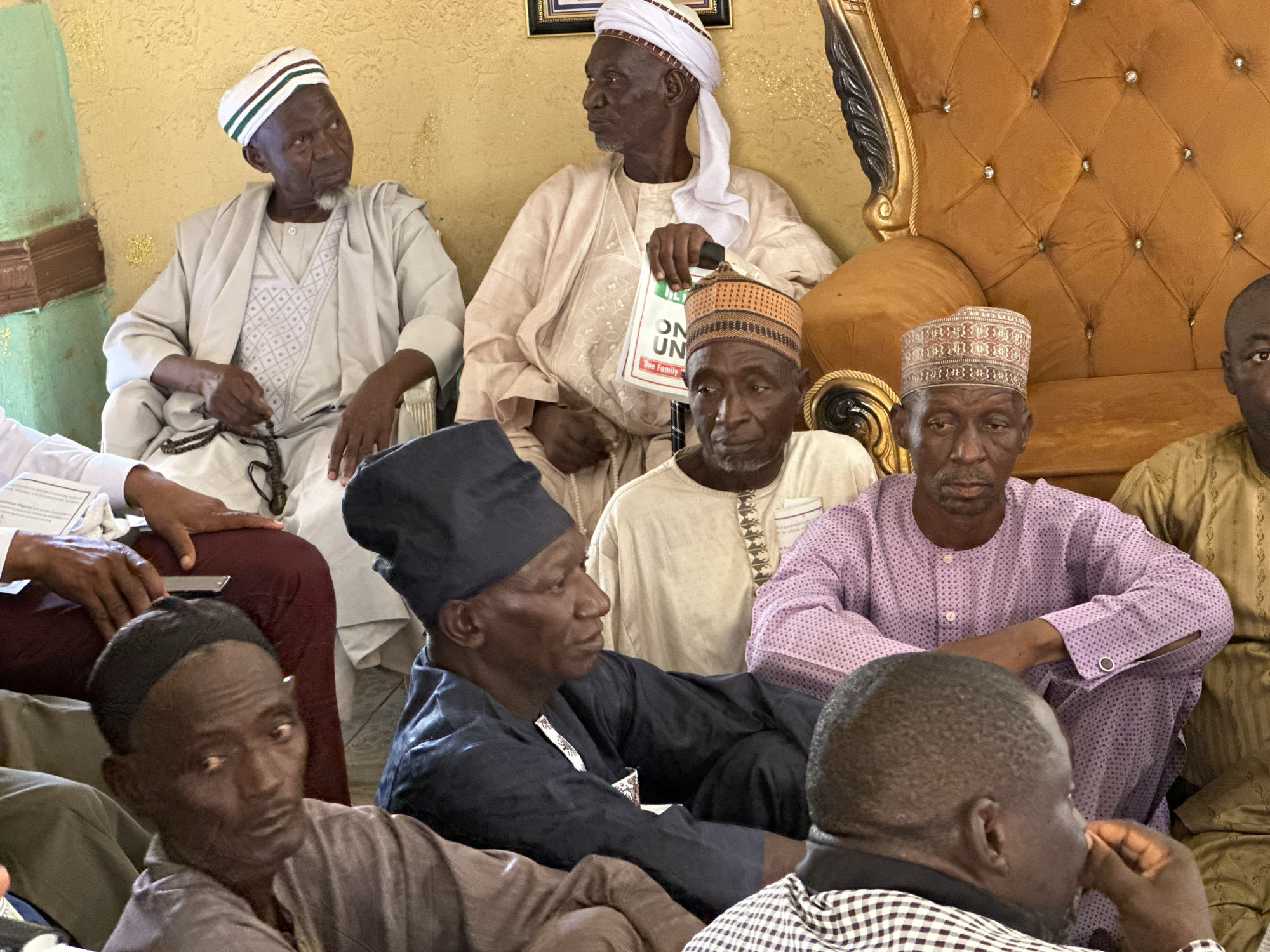 The image shows a group of older men in a room, possibly in Unknown, Unknown. Many are wearing traditional clothing and headwear associated with certain African cultures. The men are seated, some are resting their hands on their knees or holding objects. There is an ornate chair visible in the background and signs of some political affiliation are present in the form of an election advertisement in one of the subject's hands.