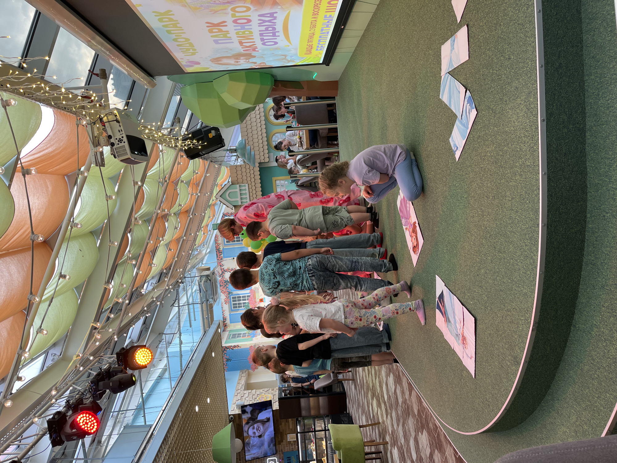 A wide shot shows a group of about 10 children participating in an activity on a stage in an indoor area, possibly a shopping mall, in the city of Unknown, Unknown. The children are lined up and appear to be looking at or interacting with pieces of a large image laid out on the green carpet of the stage.
The stage is decorated with cartoonish elements, including stylized buildings and trees. Behind the children, a screen displays an image in Russian.
Above the stage, a complex array of lighting and decorative lights is suspended from the ceiling. The ceiling itself is high and partly transparent, allowing natural light to filter in. In the foreground, to the right, a portion of seating can be seen.