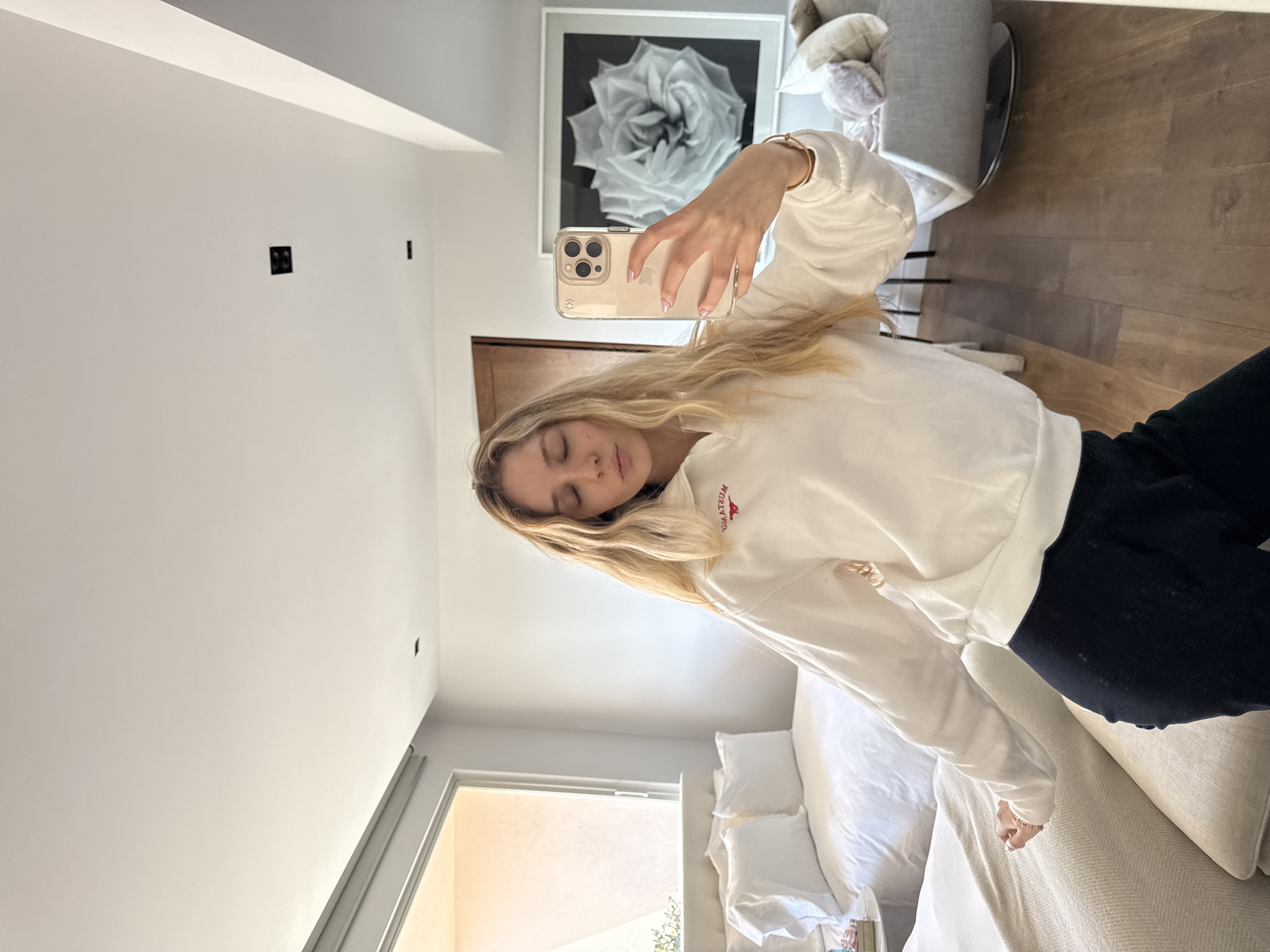 A young woman is captured taking a mirror selfie in what appears to be a modern bedroom or living space in Mexico City, Mexico. She is shown from the waist up, with her long, wavy blonde hair falling across her shoulders and chest. She wears a casual white long-sleeved sweatshirt with the reversed text "MUSTANGS" visible on the chest, and dark pants.
Her right arm is extended, holding a gold-colored iPhone with a clear case, its triple camera lenses and Apple logo clearly visible as she photographs herself. Her eyes are closed or cast downwards, giving her a relaxed and casual expression.
The background reveals a brightly lit room, likely during the daytime, with natural light streaming in from a window in the lower left portion of the image. The interior features pristine white walls and warm-toned wooden flooring. A large, framed black and white photograph of a rose hangs on the wall above her shoulder, reflecting in the mirror. A modern gray upholstered chair or ottoman is partially visible to her right. A neatly made bed with white linens and multiple pillows fills the lower part of the scene, suggesting a comfortable and contemporary home environment.