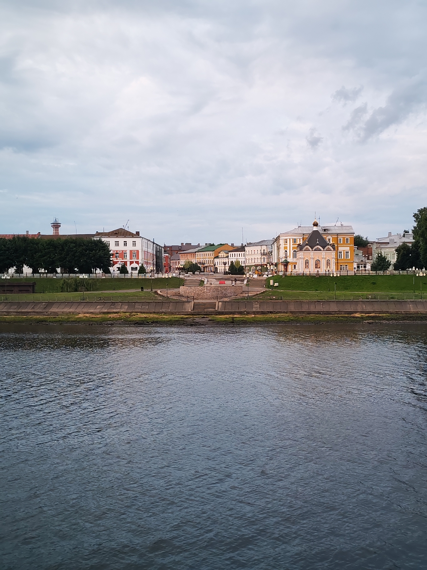The image depicts a wide, calm river in the foreground, reflecting the overcast sky. On the far bank, a picturesque cityscape lines the riverfront. Numerous buildings of varying architectural styles and colors are visible, including structures with red and white facades, yellow buildings, and some with green roofs. One notable building features a golden dome, characteristic of Russian Orthodox churches. A manicured green lawn runs along the riverbank, separated from the water by a concrete embankment. Steps and pathways are integrated into the embankment, suggesting public access to the waterfront. Trees with lush green foliage are interspersed throughout the cityscape and along the river's edge. The overall atmosphere is serene and architectural, showcasing a typical urban waterfront scene in Russia.