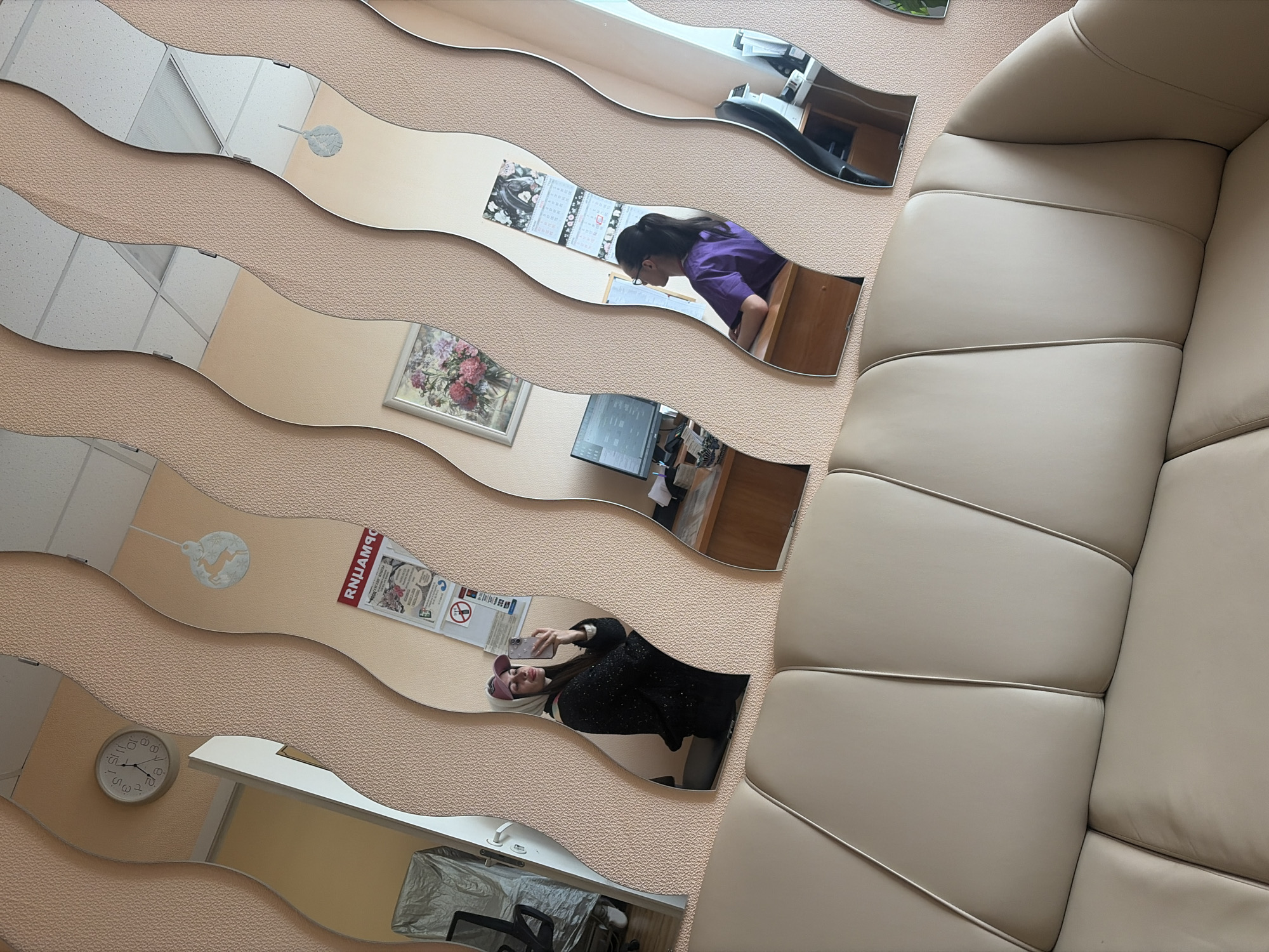 The image depicts a room with wavy, decorative mirrors on a light peach-colored textured wall. The mirrors reflect different areas of what appears to be an office or waiting room.
From top to bottom, the mirrors reflect the following:
* A ceiling with a square grid pattern (possibly a drop ceiling).
* A woman with dark hair and glasses in a purple shirt, sitting at a desk in front of a calendar.
* A framed picture (possibly of flowers) and a computer monitor on a desk.
* A Christmas ornament or decoration, and a posted sign in Russian.
* A woman with a burgundy cap taking a selfie with a phone.
* A chair and a covered object (possibly medical equipment).
A large, beige, segmented sofa occupies the right side of the image, adjacent to the wavy mirrors.
Based on the sign in Russian, the location may be Saint Petersburg, Russia. The environment appears to be a professional setting, such as a medical or dental office.