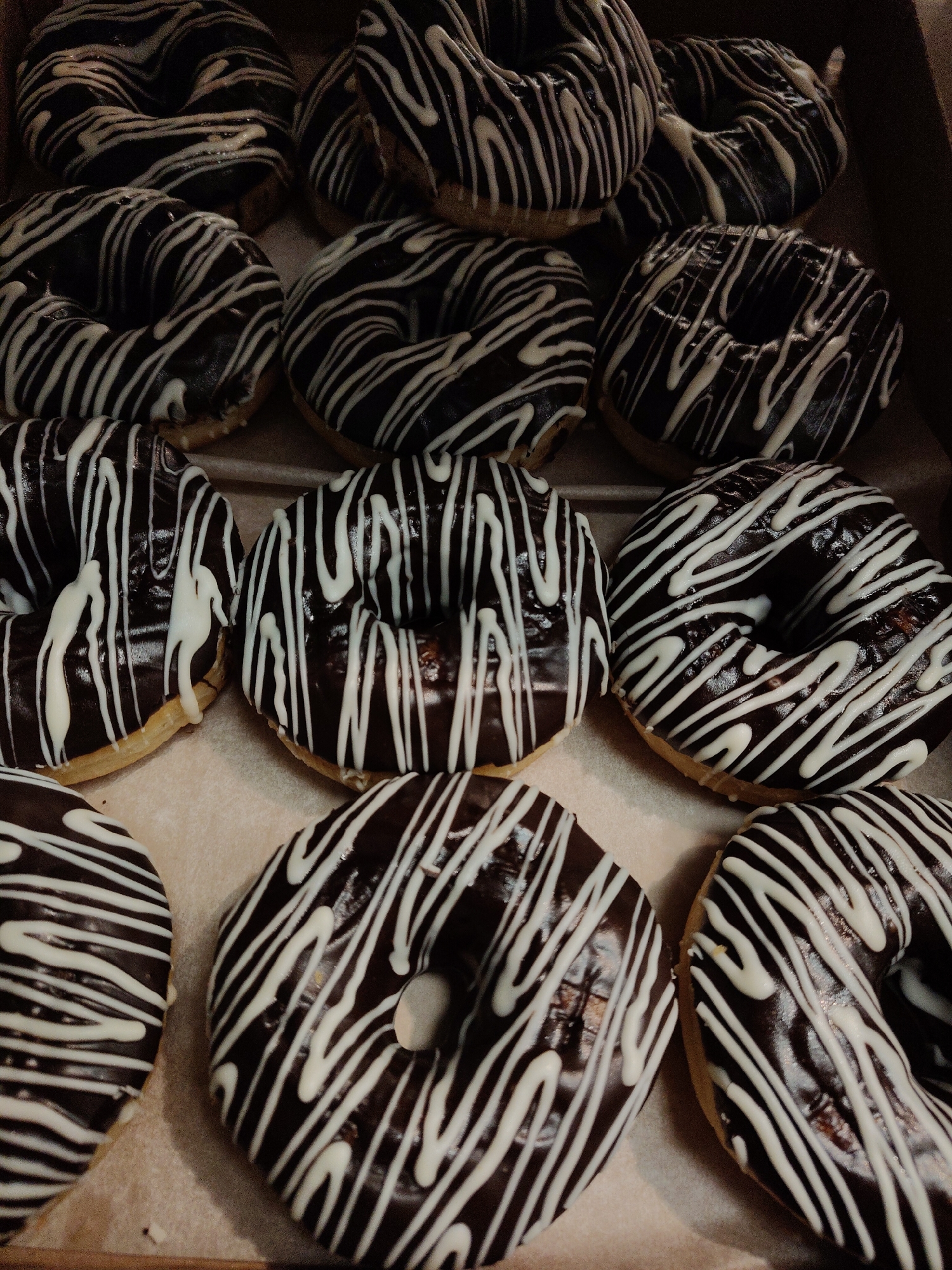 The image shows a close-up of a box of doughnuts. The doughnuts are glazed with dark chocolate and drizzled with white icing in a striped pattern. The background is a tan color, likely the interior of the box. The location is unknown.