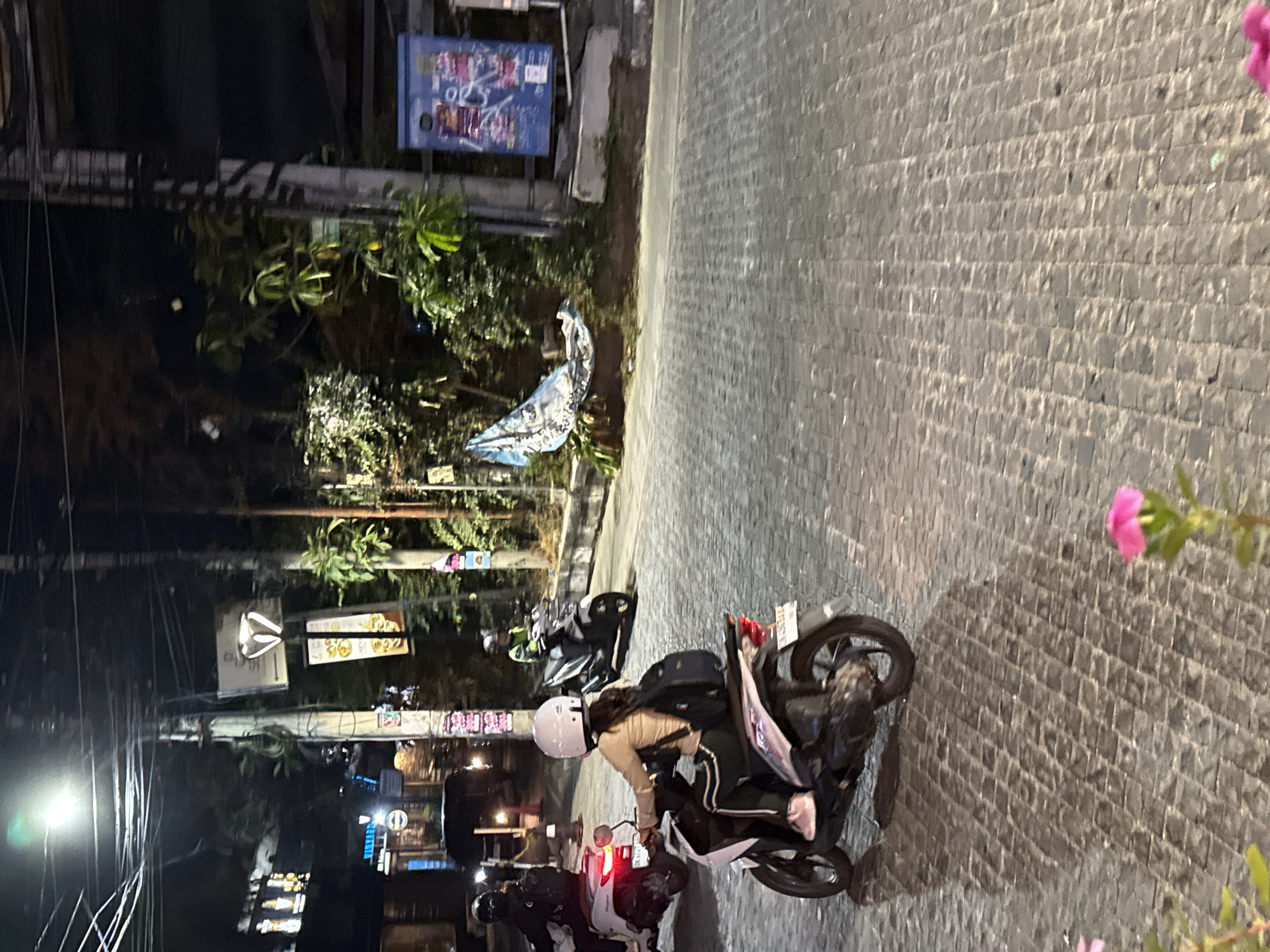 In Denpasar, Indonesia, at night, a street scene unfolds with a cobblestone pavement. Several individuals are riding scooters. In the foreground, a person wearing a light brown jacket, dark pants with green stripes, and a white helmet rides a white scooter. This scooter has a pink rear light and a license plate. Behind this scooter, another scooter is visible, also being ridden by an individual in a helmet. Further in the background, more scooters and people are present. The environment is characterized by streetlights illuminating the scene, power lines overhead, and various plants. A blue electrical box with posters is also visible on the right side of the image. A patterned cloth or flag is draped over some foliage on the left.