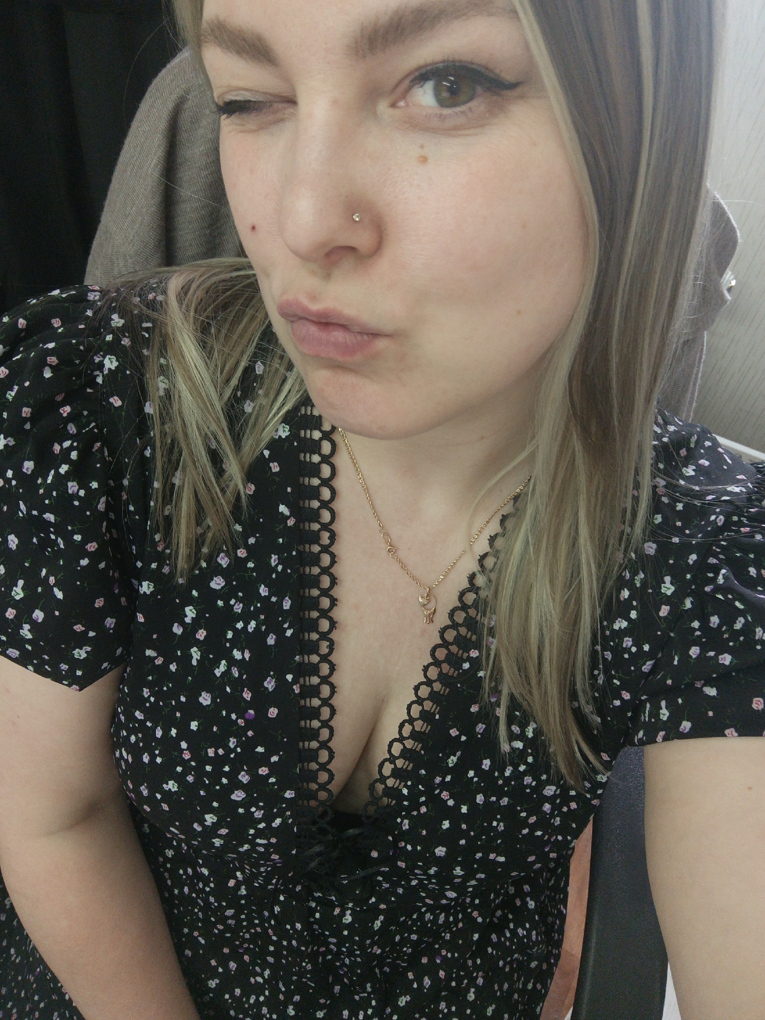 A woman is facing the camera and winking. She has fair skin and blonde hair with darker streaks. She has a nose piercing and is wearing a black floral dress with a decorative scalloped neckline and a gold necklace with a small pendant. The background is out of focus, but suggests an interior space. No information suggests a location in Noviy, Russia.