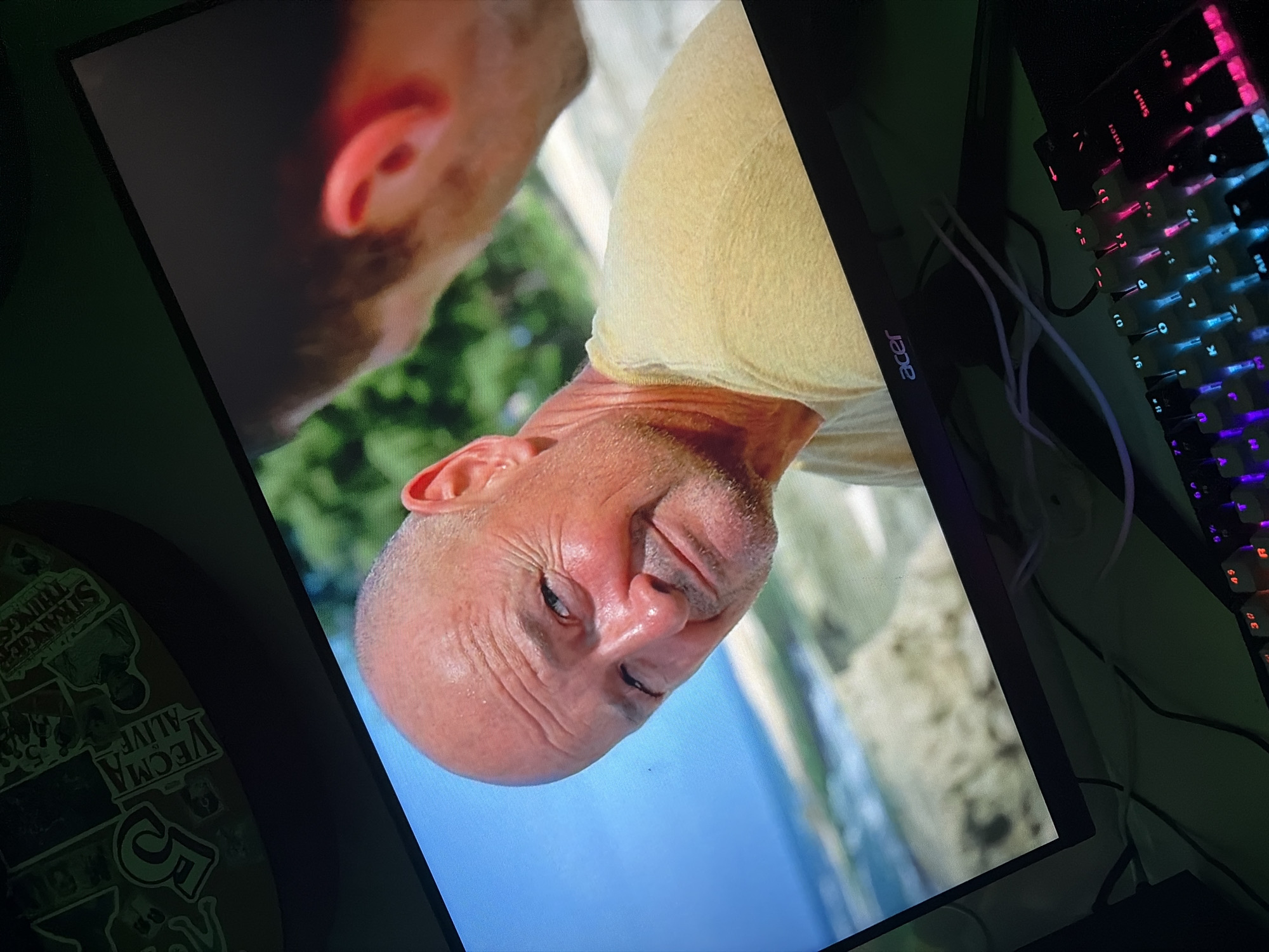 A close-up shot shows a bald, middle-aged man with weathered skin, looking slightly to his left with a faint smile. He is wearing a light yellow t-shirt. Behind him, another person's head and shoulder are visible, out of focus. The background appears to be a beach scene with blue sky and sand. The image is displayed on a computer monitor, with a keyboard visible in the foreground, lit by colorful LEDs. The monitor has the "acer" logo on its bottom bezel. The surrounding environment is dark, suggesting it is nighttime or a dimly lit room.