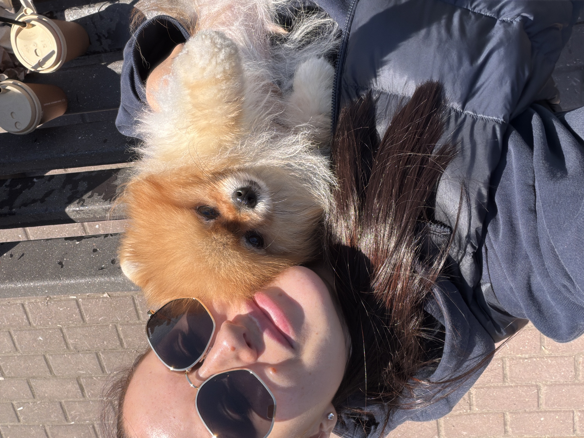 A woman with dark brown hair is lying on her back, wearing sunglasses. A Pomeranian dog is lying on her chest, partially obscuring her face. She is wearing a blue jacket and shirt. Two coffee cups are visible to the left. The ground appears to be brick. The location is Khimki, Russia.