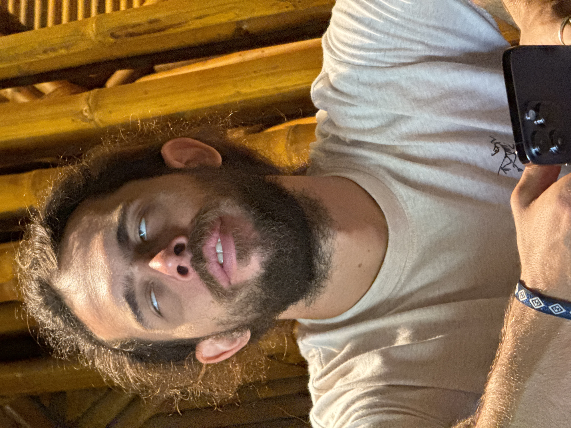 The image, set in Mù Cang Chải, Vietnam, captures a male individual from the chest up, positioned against a backdrop of vertical yellowish-orange bamboo poles. He possesses dark, curly, shoulder-length hair and a full, dark beard and mustache. His facial expression is distinctive, with his eyes rolled upwards, revealing primarily the whites (sclera) and a sliver of blue iris. His mouth is slightly open, with the upper lip slightly raised, showing his upper teeth.
He wears a light beige, short-sleeved t-shirt with a subtle texture, featuring a small, dark, stylized animal logo (resembling the Arc'teryx emblem) on the left chest area. His forearms are visible, appearing tan and hairy. On his left wrist, he wears a blue fabric bracelet with repeating white diamond-like patterns. His right hand holds a dark-colored smartphone, seen from the rear, prominently displaying a multi-lens camera module. The lighting is strong and directional, illuminating the man's forehead and casting shadows under his eyes and chin. The bamboo poles in the background are arranged vertically, creating a textured, natural wall or screen.