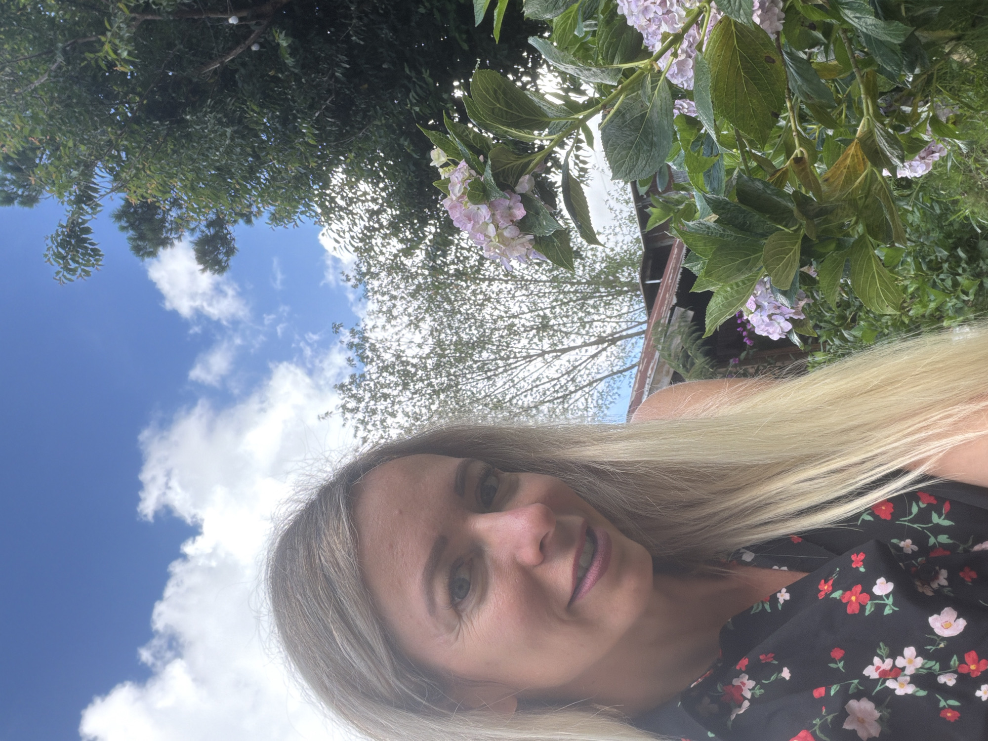 In an outdoor setting with a cloudy blue sky, a woman with long blonde hair is taking a selfie. She is wearing a black shirt with a floral pattern of red and pink flowers. She is positioned on the left side of the frame, with her face tilted towards the camera. To her right, a bush of light pink hydrangeas is in bloom. In the background, there are trees and a rustic structure with a corrugated metal roof, suggestive of a rural or semi-rural environment, potentially near Xuân Thành, Vietnam.