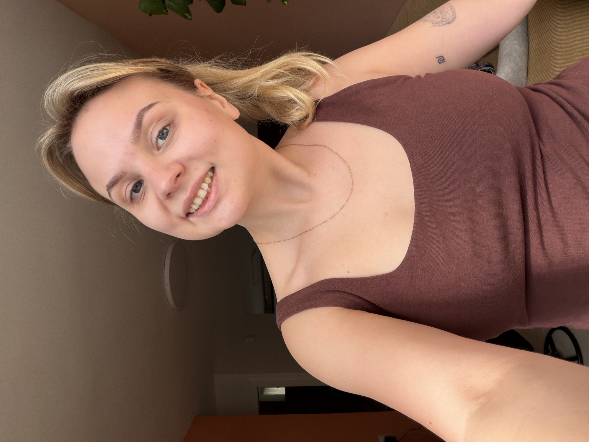 One individual, identified as a female with fair skin, blonde hair styled in a ponytail, and blue eyes, is depicted in a close-up selfie. She is smiling, revealing her upper teeth, and looking directly into the camera. She wears a brown, square-neck tank top and a thin gold-colored chain necklace. Visible on her left bicep (viewer's right arm) are two tattoos: a black outline of the Israeli New Shekel (₪) symbol and a faded, abstract circular design. The subject is indoors, illuminated by natural light entering from the right, casting parts of the scene in shadow. The background features light brown/beige walls and ceiling, with a segment of orange wall on the far left. A round white ceiling light fixture is partially visible above and behind her head. A dark green plant is partially visible on the far right, and a light brown sofa or similar furniture item is behind her on the right. A dark doorway or opening is visible in the distant left background. Based on the presence of the Israeli New Shekel symbol tattoo, the scene is situated within the context of Israel, specifically referenced as Tel-Aviv per the request.