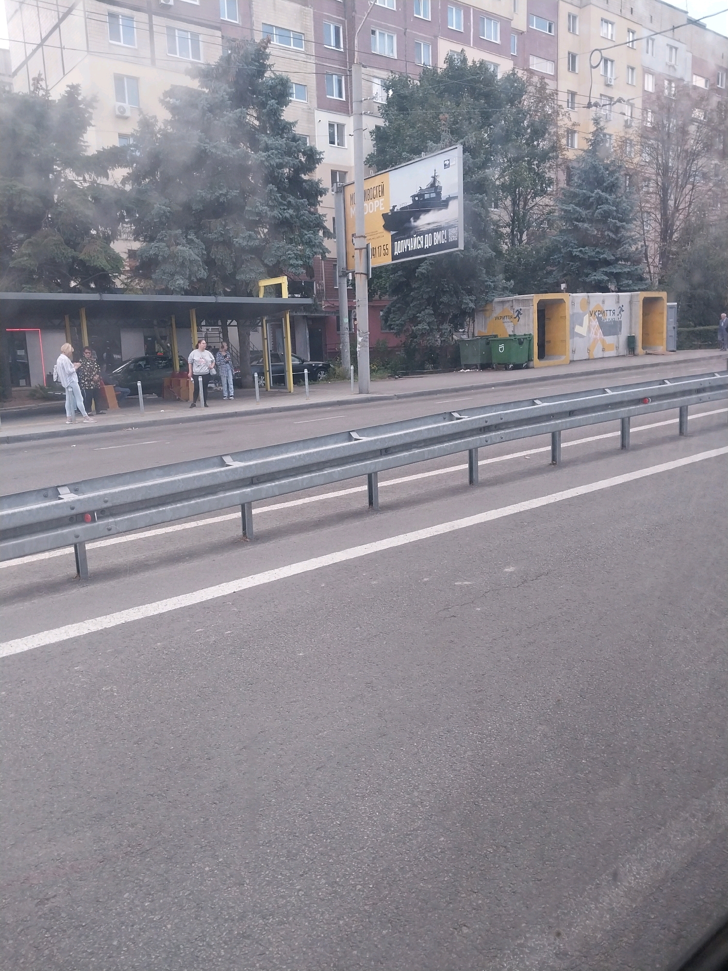 A medium shot captures a scene on a street in Dnipro, Ukraine, on a cloudy day. In the foreground, a metal guardrail runs parallel to a paved road with a white stripe marking. Behind the guardrail, a bus stop shelter with yellow supports covers a sidewalk where several people are standing. One person, a woman in a striped shirt and light-colored pants, stands closest to the camera. Another woman, in a grey t-shirt and jeans, is looking to her right. Two men are also present, one in a patterned shirt and another in a plaid shirt. Behind the bus stop, several trees with dark green foliage rise up. A billboard advertising a boat and text in Cyrillic is visible above the trees. To the right of the bus stop, two large green trash bins are situated next to a concrete structure with arched entrances, also adorned with Cyrillic text and yellow accents. In the background, a multi-story apartment building with numerous windows is visible. The overall atmosphere is mundane, depicting a typical urban street scene.