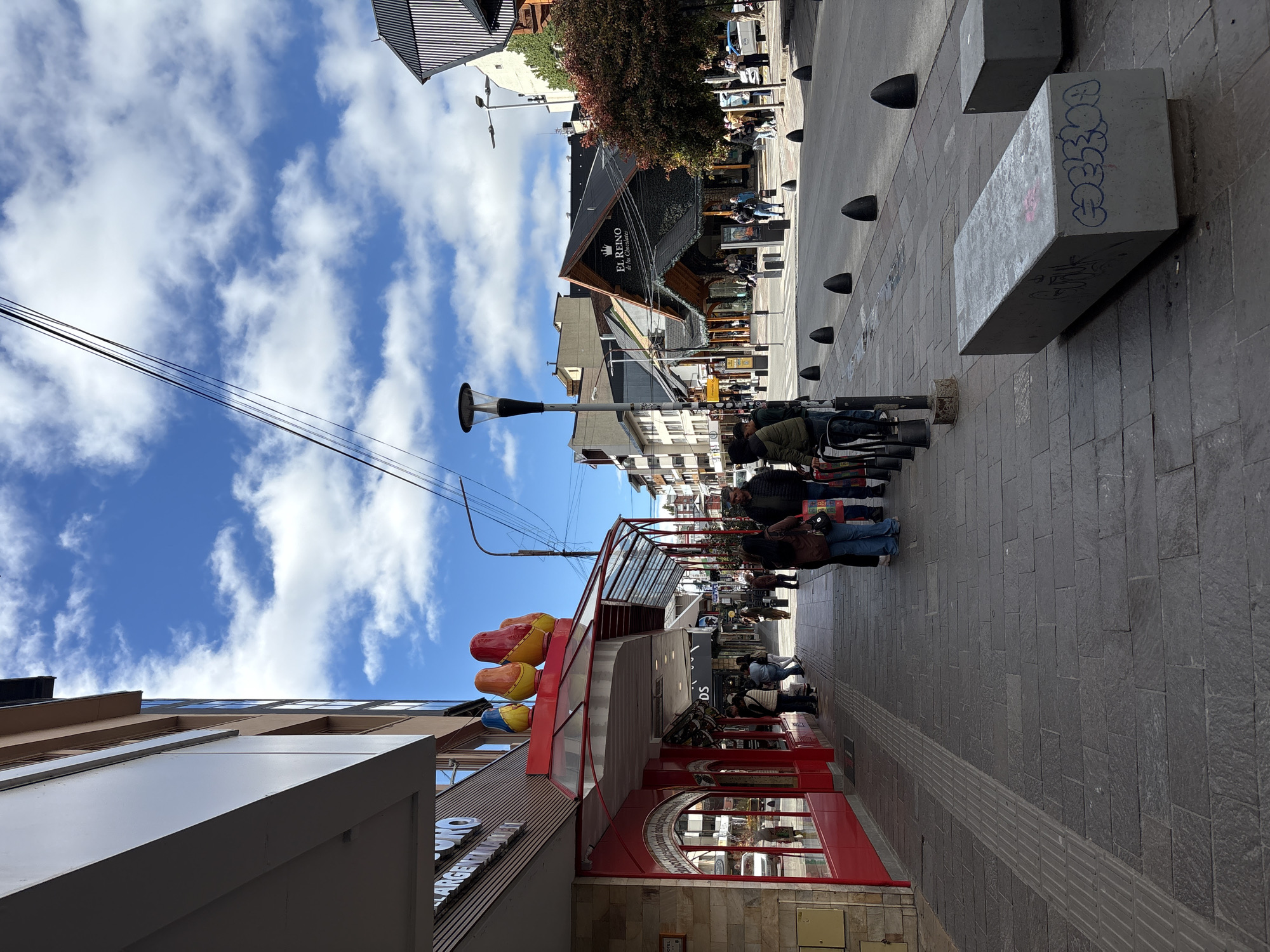 A slightly angled view of a pedestrian street in San Carlos de Bariloche, Argentina, with a bright blue sky and scattered clouds above. The street is paved with gray rectangular stones. On the right side, a row of dark, rounded bollards separates the pedestrian area from the street. Concrete benches with graffiti are also visible. Several people are walking along the street, dressed in casual clothing, some in jackets and jeans.
On the left, the corner of a building is visible, with a red-framed storefront adorned with colorful balloons. A store sign with some lettering can be seen. Further down the street, buildings with dark, pitched roofs can be seen, including "EL REINO de los Chocolate". Streetlights are present along the thoroughfare, and wires extend across the sky.