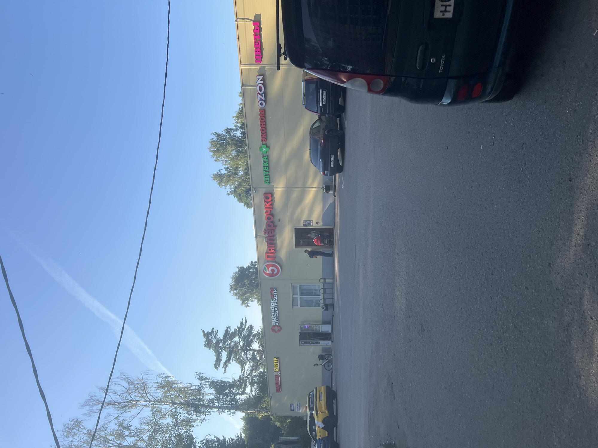 The image shows the exterior of a strip mall in Pushkino, Russia, on a clear, sunny day. The building facade is light yellow with various signs in Cyrillic. From left to right, the signs indicate a burger shop, "autodoc auto parts," a "Pyaterochka" grocery store, an "Apteka" (pharmacy), "Econom," "OZON," and "Flowers."
In the foreground, a dark green Toyota minivan is parked, partially obscuring a black SUV. To the left of the minivan, a yellow taxi is visible. In front of the strip mall, several people are present. Two individuals, one with a stroller, are near the entrance of the Pyaterochka store. Another person stands near the entrance of the autodoc store, and a bicycle is parked outside. The scene takes place on an asphalt parking lot. In the background, trees and a bright blue sky with contrails from an aircraft are visible. Overhead power lines are also present.