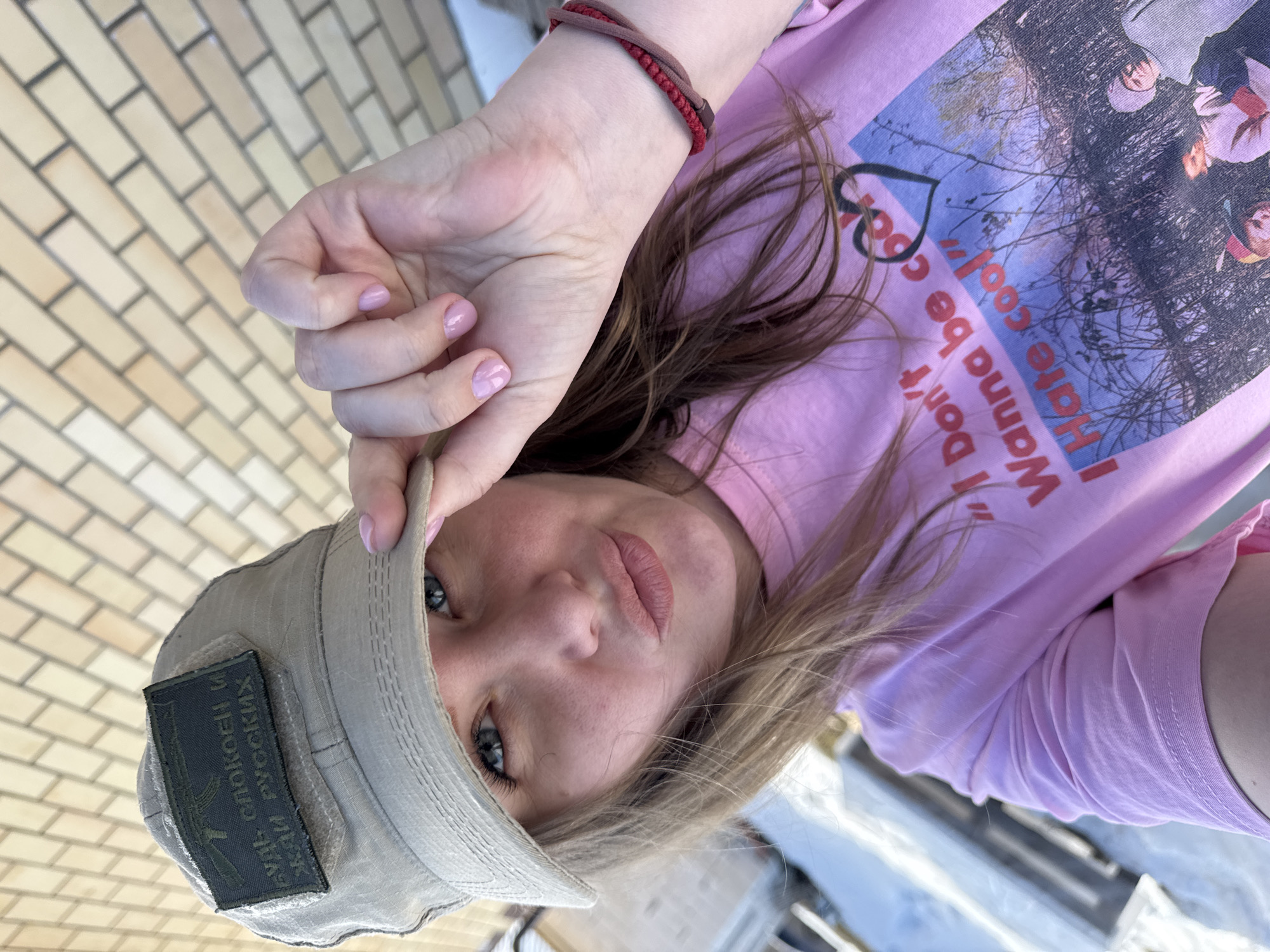 A female individual is depicted in a close-up, head-and-shoulders selfie taken in Gornyi (Dvurechenskoe S/P), Russia. She possesses light-colored hair and blue eyes, and her lips are slightly puckered. Her left hand, adorned with light pink nail polish, holds the brim of a light khaki-colored, boonie-style cap. Affixed to the cap is a dark green rectangular patch featuring white Cyrillic text, "БУДЬ СПОКОЕН И ЖДИ РУССКИХ" (Be calm and wait for the Russians), accompanied by an illustration of an AK-style rifle. She wears a pink long-sleeved t-shirt with a graphic design that includes the English text "I Don't Wanna be cool I Hate cool!" and an image of several individuals outdoors. A red and brown braided band is visible on her right wrist. The immediate background behind her head is a light-colored brick pattern. Blurry outdoor elements are visible to the left of the frame.