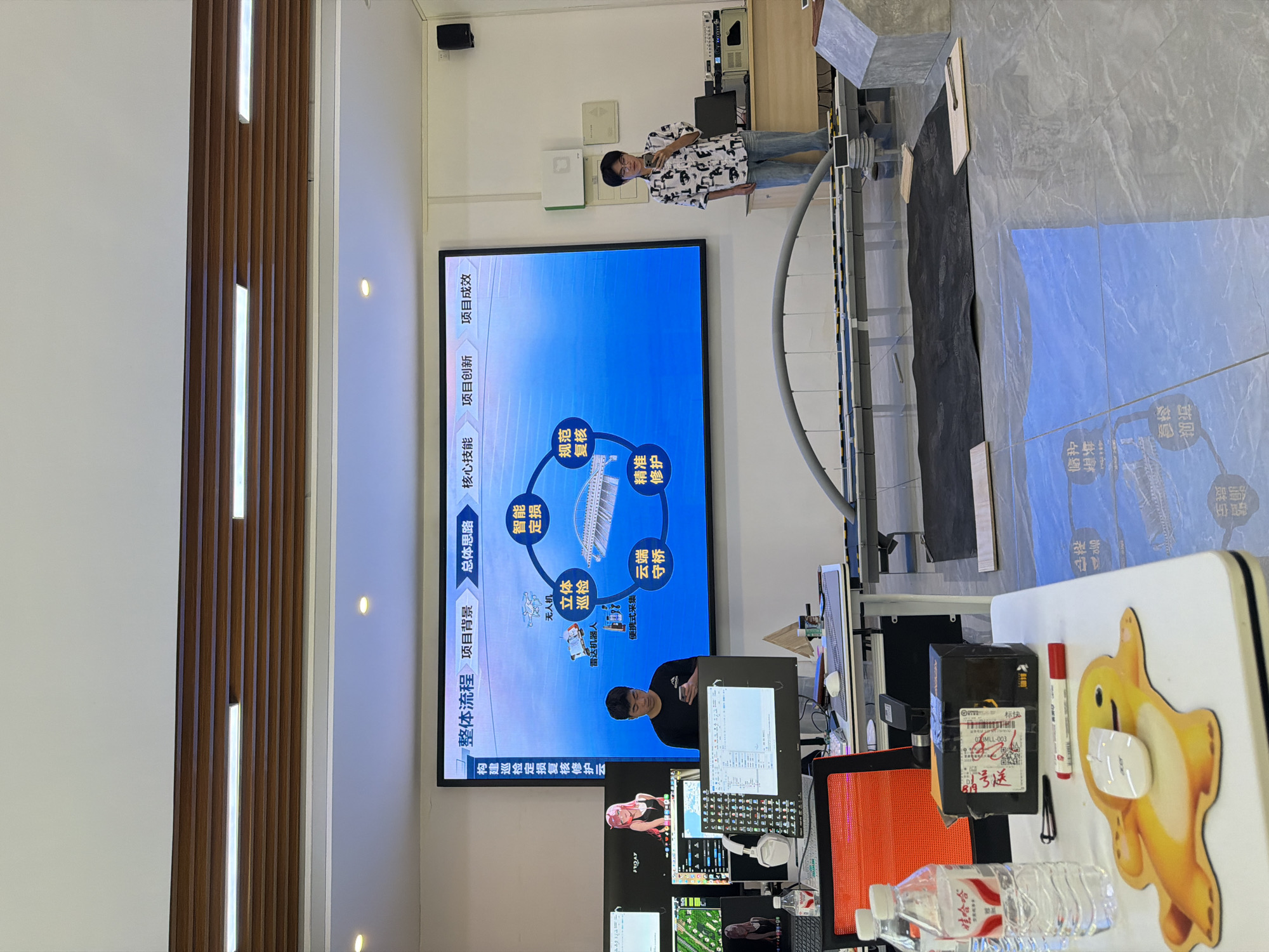 The image depicts a presentation room, possibly in Jiangjin, China, with a large digital display screen showing a flowchart for a project. The screen displays text in Chinese, including "整体流程" (Overall Process), "项目背景" (Project Background), "总体思路" (Overall Idea), "核心技能" (Core Skills), "项目创新" (Project Innovation), and "项目成效" (Project Effectiveness). Several nodes within the flowchart include terms like "无人机" (Drone), "立体巡检" (Stereoscopic Inspection), "雷达机器人" (Radar Robot), "便捷式采集" (Convenient Collection), "智能定损" (Intelligent Damage Assessment), "规范复核" (Standardized Review), and "精准修复" (Precise Repair).
In the foreground, a young man in a black t-shirt is standing behind a desk, looking at his phone. In front of him is a computer monitor displaying what appears to be a program with code. To his right, another young man in a patterned shirt and jeans is holding a phone and speaking into a microphone, facing the screen.
The room contains multiple computer monitors, headphones, a water bottle, and office supplies on desks. In the center of the room, a model of a bridge structure is set up on the floor, with black and blue mats laid out around it. The walls are white, and a wooden ceiling with integrated lighting is visible. The floor is tiled with a marble-like pattern. The overall environment suggests a technology or engineering-related presentation or workspace.