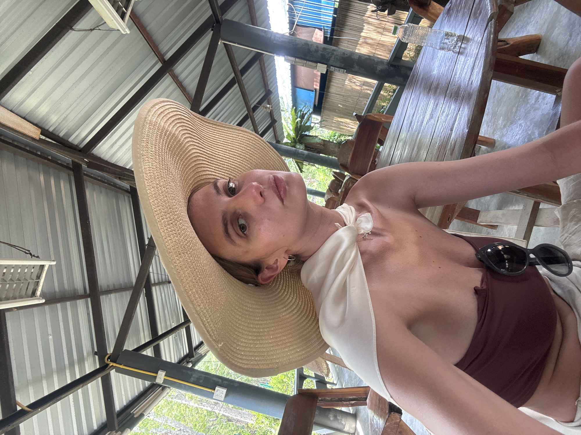 A sideways eye-level shot features a woman wearing a wide-brimmed straw hat, a brown strapless top, and a white scarf tied around her neck. Sunglasses are resting on her chest. She's seated at a wooden table, a plastic bottle visible on the table's surface. The setting seems to be an outdoor or semi-outdoor area with a corrugated metal roof supported by metal beams. Green foliage is visible in the background. The location is indicated as Unknown, Unknown.