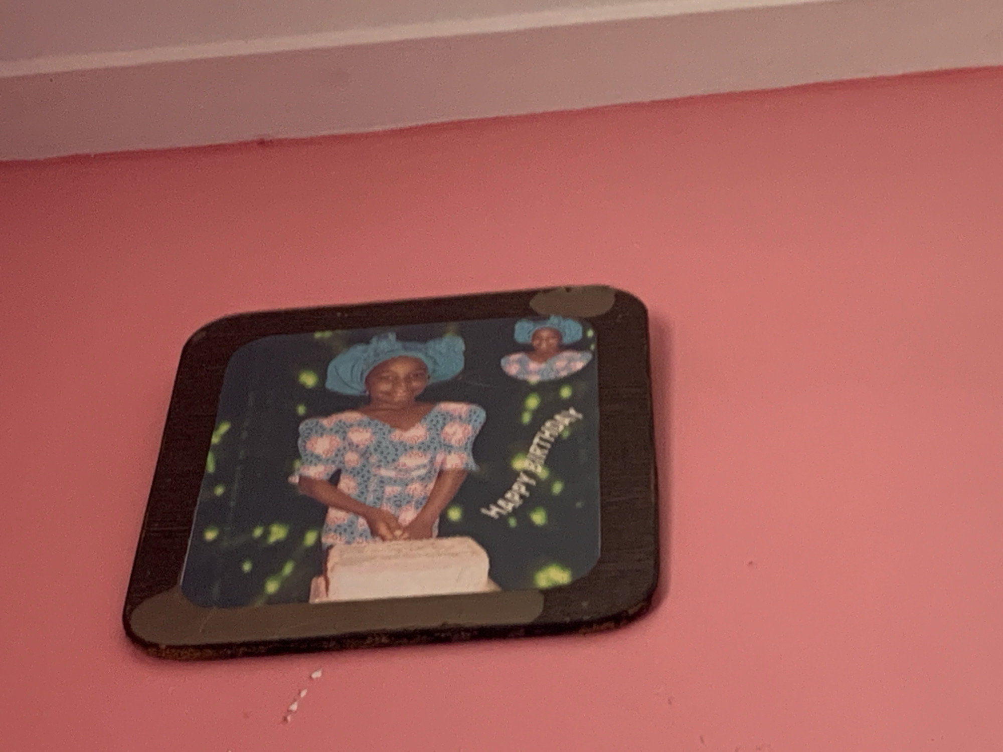 A framed photograph is displayed on a pink wall, likely in Kakau, Nigeria. The photograph features a young woman in traditional attire, including a blue headscarf and a patterned blue and pink dress. She is standing in front of a cake, with her hands positioned as if to cut it. The background of the photo is dark with scattered green and yellow confetti-like shapes. Above the main image, a smaller inset photo shows a close-up of the same woman's face, also wearing a blue headscarf. White text, curving across the photograph, reads "HAPPY BIRTHDAY." The overall impression is a celebratory and personal moment captured in time. The lighting in the original image suggests indoor illumination. The pink wall and the somewhat worn frame of the photograph suggest a lived-in domestic setting.