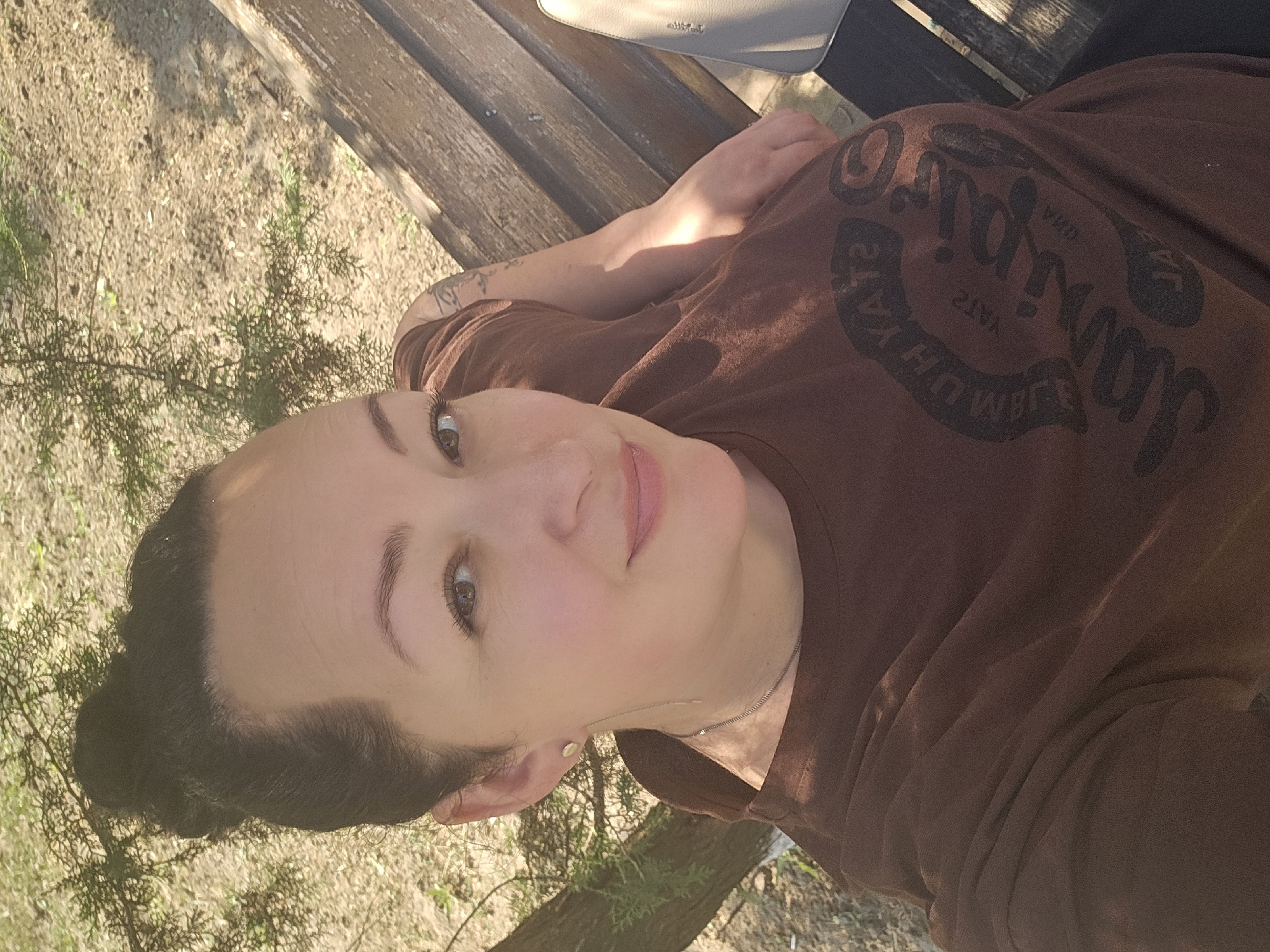 This is a close-up, horizontal, outdoor selfie of a woman. She is lying on a wooden bench, with her head tilted to the side and looking directly at the camera. She has dark hair styled in a bun and appears to be wearing a brown t-shirt with a graphic design on it. The graphic design appears to be a stylized text, but it is difficult to read clearly due to the angle and resolution.
The background shows a sandy ground with some green foliage, suggesting a park or natural setting. Sunlight is casting shadows on the bench and the woman's arm, indicating it is daytime. The overall mood of the image is relaxed and casual. There is a light-colored handbag resting on the bench behind her.
No specific activity is taking place other than the woman posing for a selfie. The image is set in Skadovsk, Ukraine, but no specific landmarks are visible to confirm this location context. The woman's expression is neutral to slightly smiling. The time of day is likely afternoon due to the strong sunlight and shadows.
