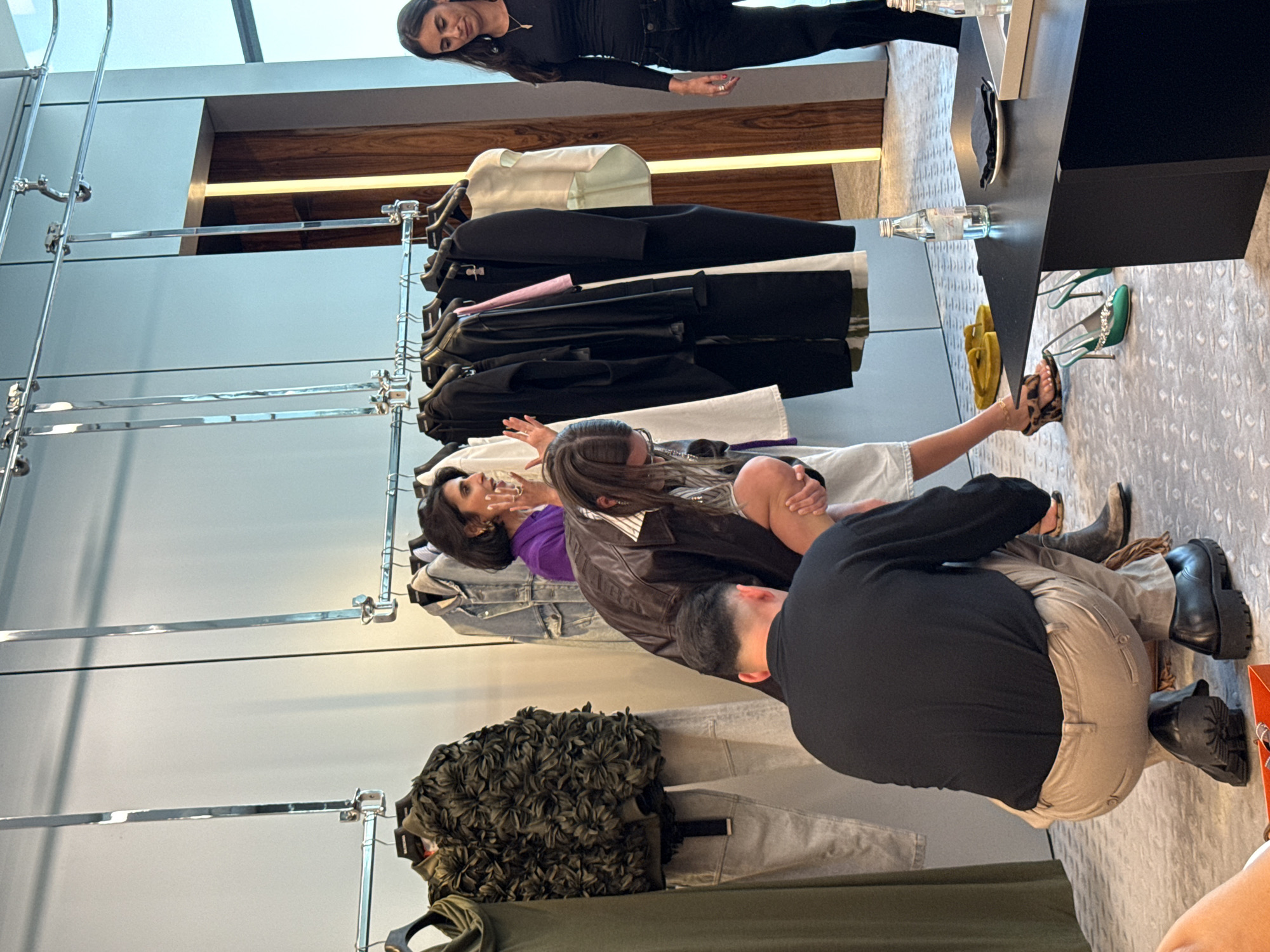 In Dubai, United Arab Emirates, within a brightly lit retail or showroom space, a group of individuals is gathered around a clothing rack and a low black table. A woman in a purple shirt gestures animatedly, engaged in conversation with a person wearing a brown leather jacket, who is examining footwear on the floor. Another person, wearing a black top and tan pants, is crouched down. To the right, a woman in a black top and jeans looks towards the table. Various garments hang on the rack, including jackets, dresses, and pants. On the table, there are bottles of water, a plate with a napkin, and a tray. The floor is covered with a patterned carpet.