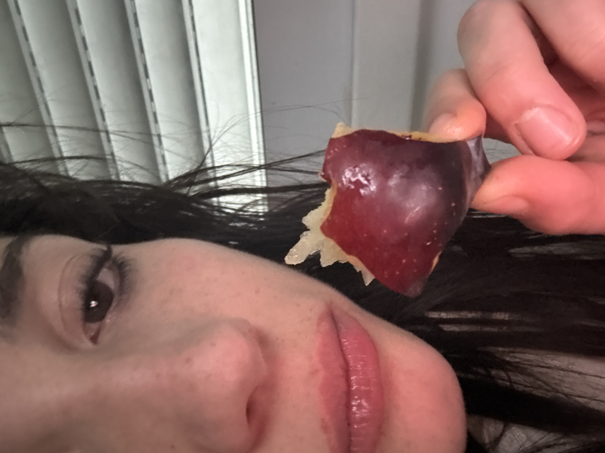 A close-up, slightly angled shot of a person holding a partially eaten fruit in their hand. The person's face is visible, featuring dark hair, an eye, a nose, and lips. The skin tone is light. The fruit is dark red on the outside and has a lighter-colored pulp exposed where it has been bitten. A hand with light skin tone and short nails is holding the fruit. In the background, vertical blinds are visible. There is no discernible information in the image to indicate that it was taken in Buenos Aires, Argentina.