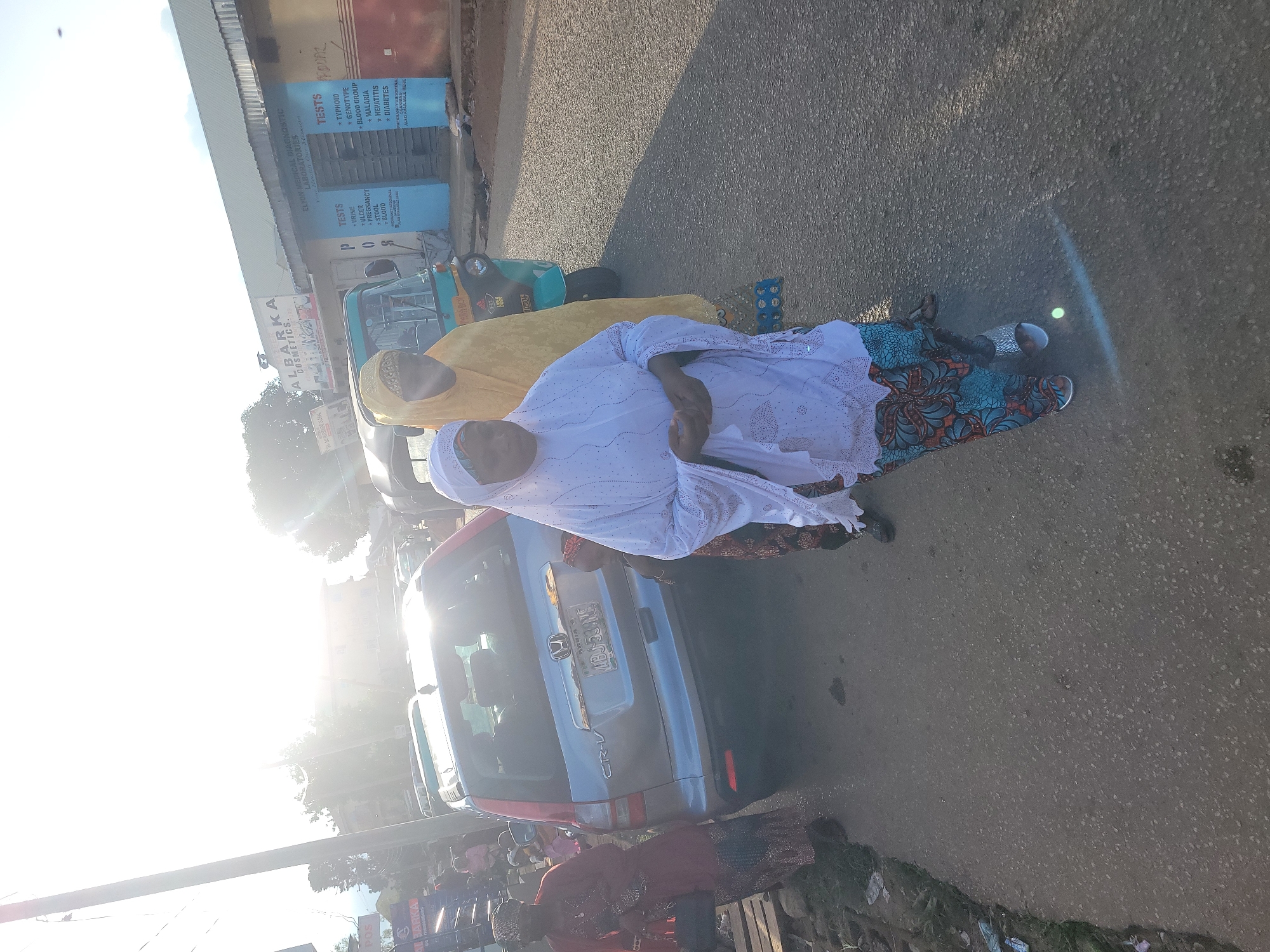 This image captures a daytime street scene in what is described as the city of Jos, Nigeria, characterized by strong sunlight and activity.
In the foreground and center, a woman is seen from the chest down, wearing a white, long-sleeved tunic adorned with perforated patterns, and patterned trousers featuring blue, orange, and brown hues. She wears silver sandals and her hands are clasped in front of her. Directly behind her, another woman is partially visible, dressed in a yellow head covering (hijab) and matching yellow garment, facing left. To the left of these women, further back, a silver Honda CR-V SUV is parked or moving slowly, displaying a Nigerian license plate "ABJ 534 NE" indicating registration from Abuja. Adjacent to the CR-V's rear, a small individual in patterned clothing is partially visible. On the far left, a woman in a dark red top and patterned bottom garment is walking, carrying a dark bag.
Behind the women in the center, and to the right of the CR-V, a light blue/turquoise Keke Napep (tricycle taxi) is partially visible. The background features several commercial buildings. On the left, a building sign reads "ALBARKA COSMETICS." Further to the right, a building displays "ELYON MEDICAL DIAGNOSTIC LABORATORIES" with a blue wall section listing various medical "TESTS" including "URINE, ULCER, PREGNANCY, STOOL, BLOOD" on the left column, and "TYPHOID, GENOTYPE, BLOOD GROUP, MALARIA, HEPATITIS, DIABETES" on the right. Below these, "PREGNANCY ABDOMINAL SCANNING ALSO AVAILABLE HERE" is stated. A faded red wall section is visible to the right of the medical laboratory.
The ground is a paved asphalt road, with some scattered debris and vegetation visible along the left edge. The scene is heavily illuminated by the sun, causing significant lens flare and overexposure in the upper portion of the image, particularly the sky and upper-left background. A utility pole is visible on the left side of the road. Shadows suggest the sun is high and slightly behind the viewer.