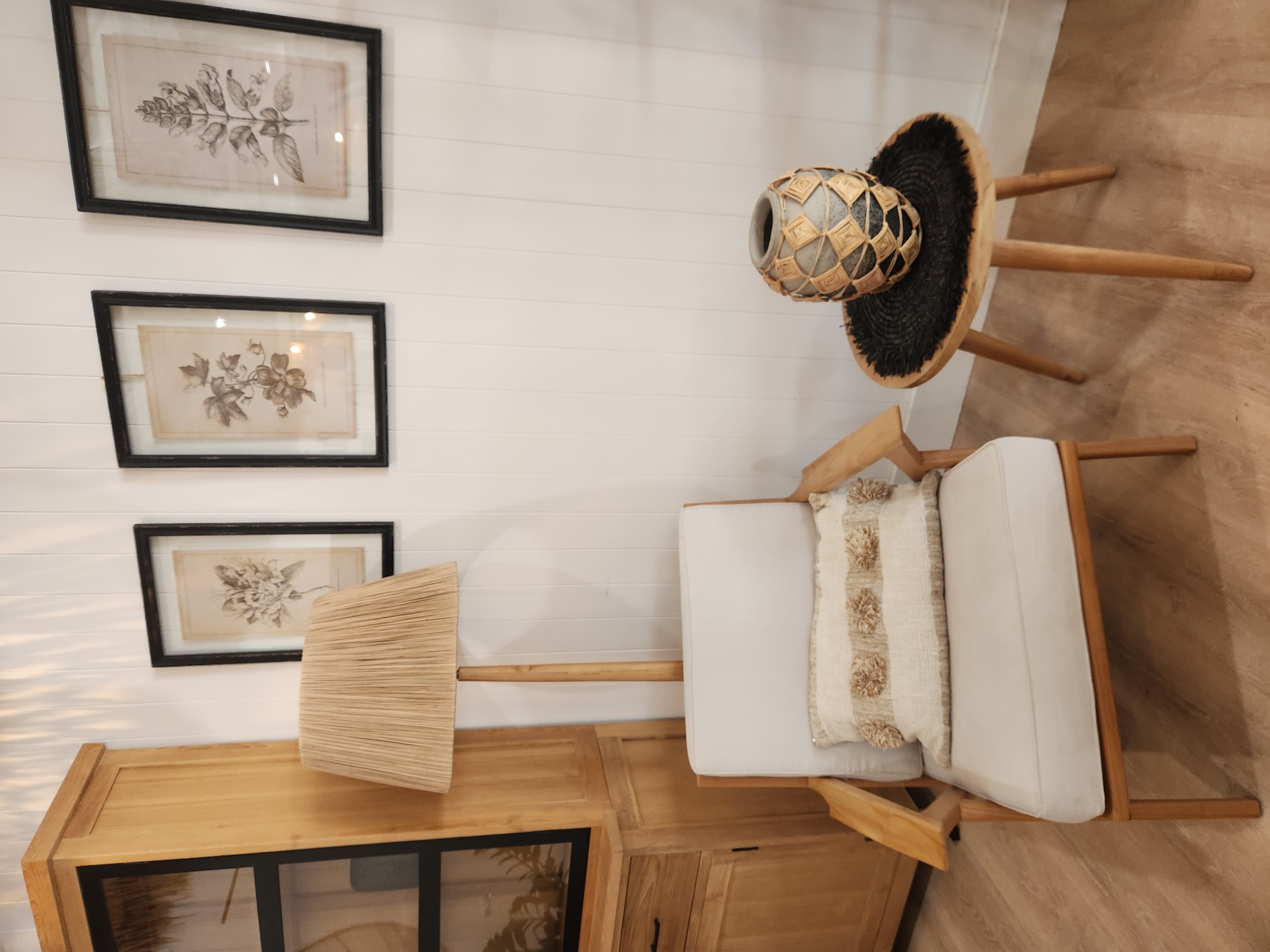 The image depicts an indoor scene with natural light. In the foreground, a light-colored armchair with wooden armrests and legs is visible. A textured cream-colored pillow with darker pom-pom accents is placed on the seat of the chair. To the right of the armchair, a small, round wooden side table with three tapered legs stands. On the table, a dark, circular woven mat supports a decorative vase. The vase is gray with an intricate pattern of woven straw-like material in diamond shapes.
Behind the armchair and side table, a floor lamp with a wide, pleated beige lampshade is positioned. To the left of the lamp, a tall wooden cabinet with glass doors and wooden shelves is seen. Three framed botanical prints hang on the white paneled wall behind the armchair and lamp. The prints are in black frames and feature detailed drawings of plants on a light-colored background.
The overall style of the room appears to be natural and bohemian, with a focus on wood textures and neutral colors. No people are present in the image. The scene is set in an interior space, possibly a living room or a display area. No specific location in the city of Maitland, United States of America, can be identified from the image.