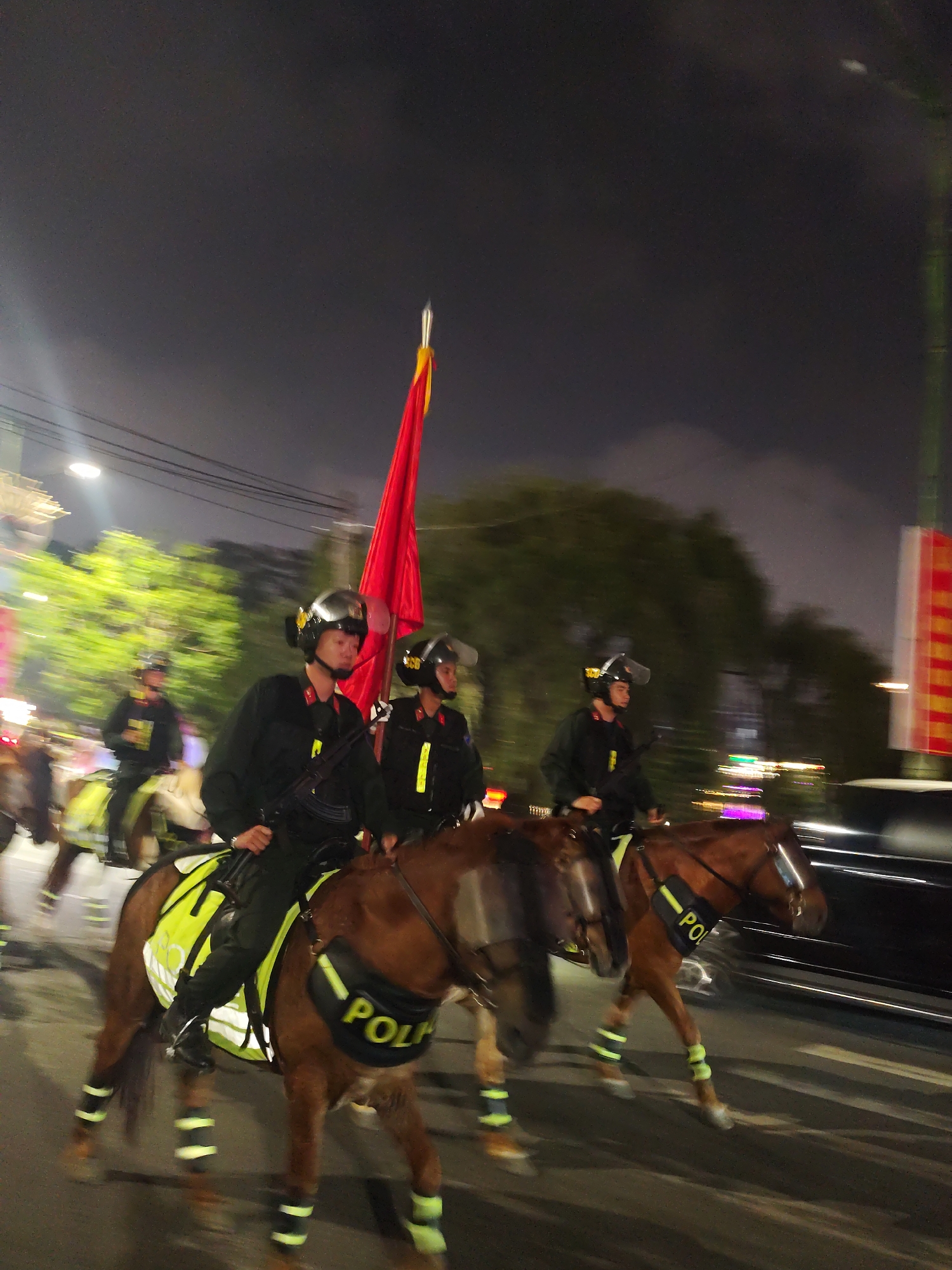 A group of police officers on horseback are participating in a nighttime parade or procession in Da Lat, Vietnam. The officers are wearing black uniforms, helmets with visors, and high-visibility yellow vests or saddle blankets. The horses are adorned with similar yellow and black accessories, including a "POLICE" band on the flank of at least one horse.
The scene appears to be a street at night, illuminated by streetlights and the headlights of vehicles. The background shows blurred trees and buildings, suggesting an urban or semi-urban setting. The overall mood is solemn and orderly, fitting for a police presence during an event. The sky is dark, indicating it is nighttime, and the weather appears clear.
One officer in the foreground carries a large red flag with a yellow symbol, possibly a national flag or a symbol related to the event. The text "POLICE" is visible on the equipment worn by the officers and their horses, confirming their role. The motion blur in the image suggests that the procession is either moving or the photograph was taken with a slight shake.