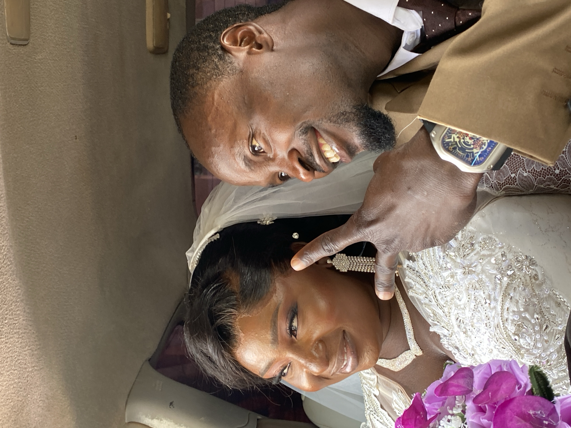 A dark-skinned man and woman, presumably a newly married couple, are indoors. The location, Kuru Karama, Nigeria, cannot be confirmed from the image. The man, wearing a brown suit and tie, smiles as he touches the woman’s face. She is wearing a white wedding dress, veil, and jewelry, and holds a purple flower bouquet. The setting appears to be inside a vehicle, based on the upholstered interior.