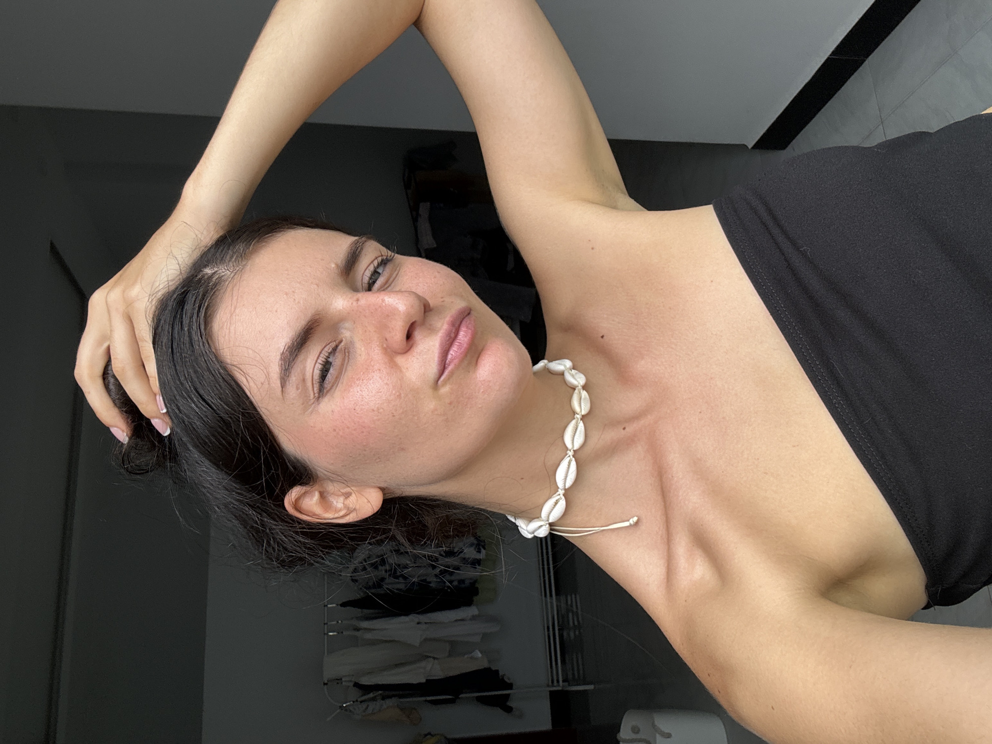 A woman is facing the camera with her arm raised above her head. She is wearing a black top and a seashell necklace. The background appears to be a room with dark-colored furniture and a window. The image provides no indication of it being located in Mũi Né, Vietnam.
