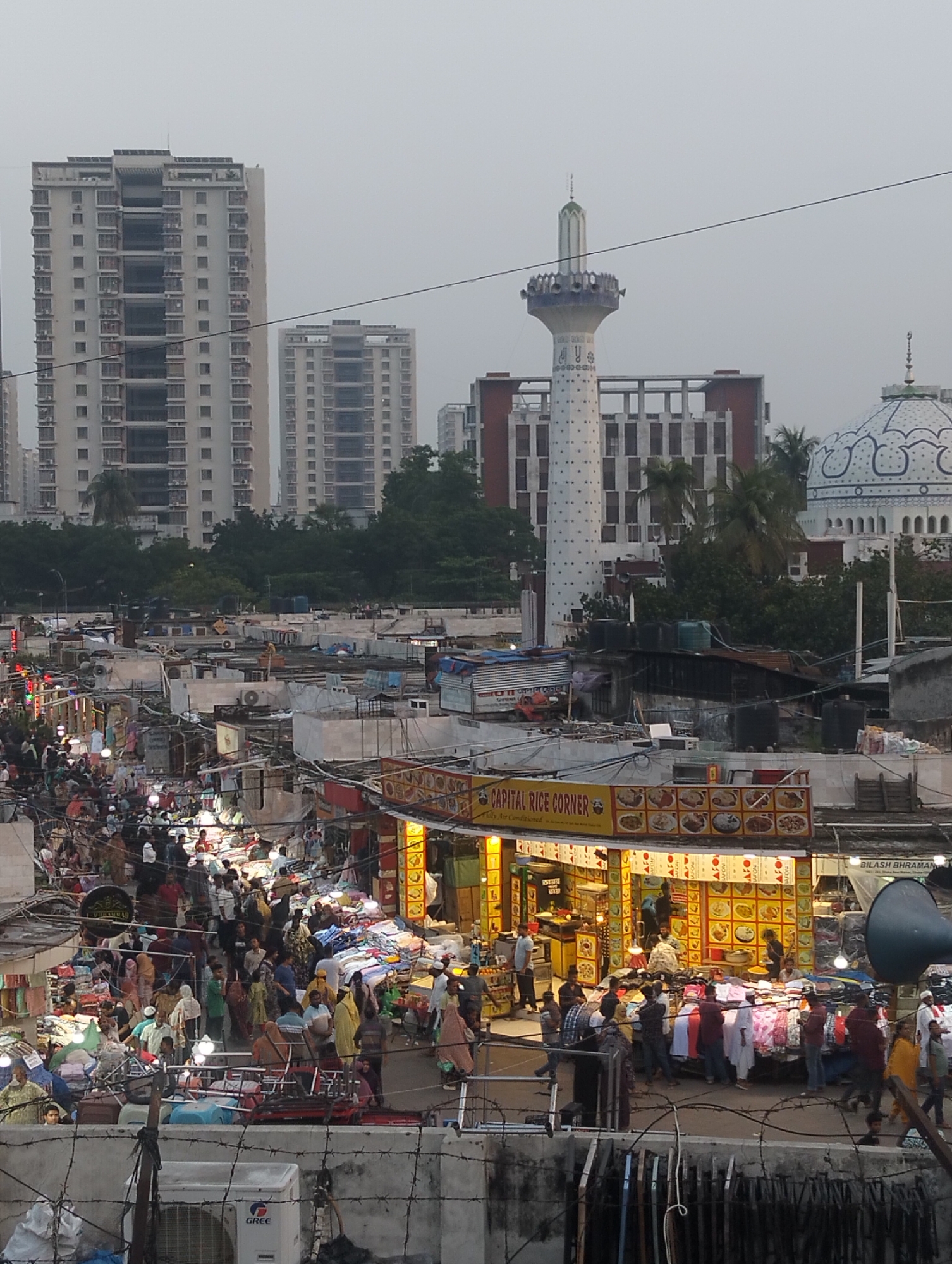 The image depicts a crowded street market in Dhaka, Bangladesh. The market, teeming with people, features numerous stalls with merchandise. Buildings, including a tall tower, a mosque with a distinctive dome, and several high-rise apartments, form the backdrop. The “Capital Rice Corner” restaurant is visible. The scene is observed from an elevated vantage point behind a fence, with air conditioning units and other debris in the foreground. The lighting suggests either dusk or dawn, with illuminated market stalls contrasting against the sky.