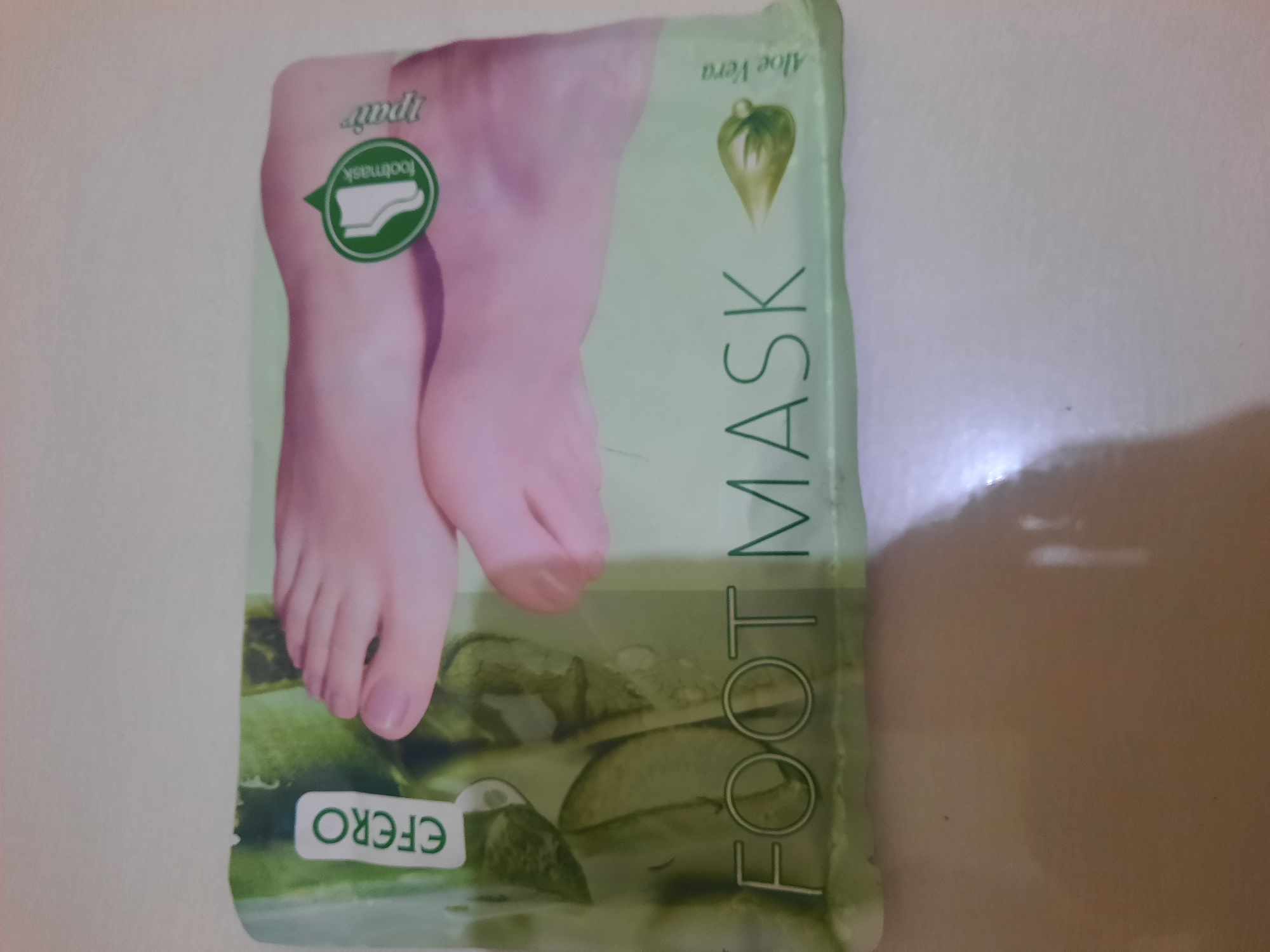 A product package for a foot mask is photographed indoors, likely on a table, against a blurred background. The package features an image of a pair of feet with pinkish skin, and text that reads "EFERO FOOT MASK" and "Aloe Vera". A circular green logo with an image of a foot inside and the text "footmask" is visible on the right side of the package, along with the word "npair". The package is oriented horizontally.