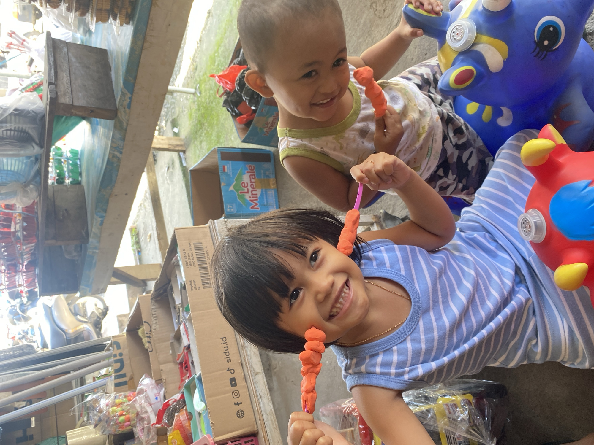 Two young children, a girl and a boy, are playing with orange clay outside in what appears to be a market stall in Ta', Indonesia. The girl, in the foreground, wears a blue and white striped sleeveless dress and a gold chain. She is smiling and holding a piece of clay shaped like a string of beads up to her cheeks. The boy, to her right, wears a white sleeveless shirt with a green trim and patterned shorts. He is also smiling and holding a piece of orange clay. They are sitting on or near colorful toy animals, one blue and one red. In the background, there are cardboard boxes, plastic bags, and market stalls, suggesting a busy commercial area. The lighting indicates it is daytime, likely sunny.