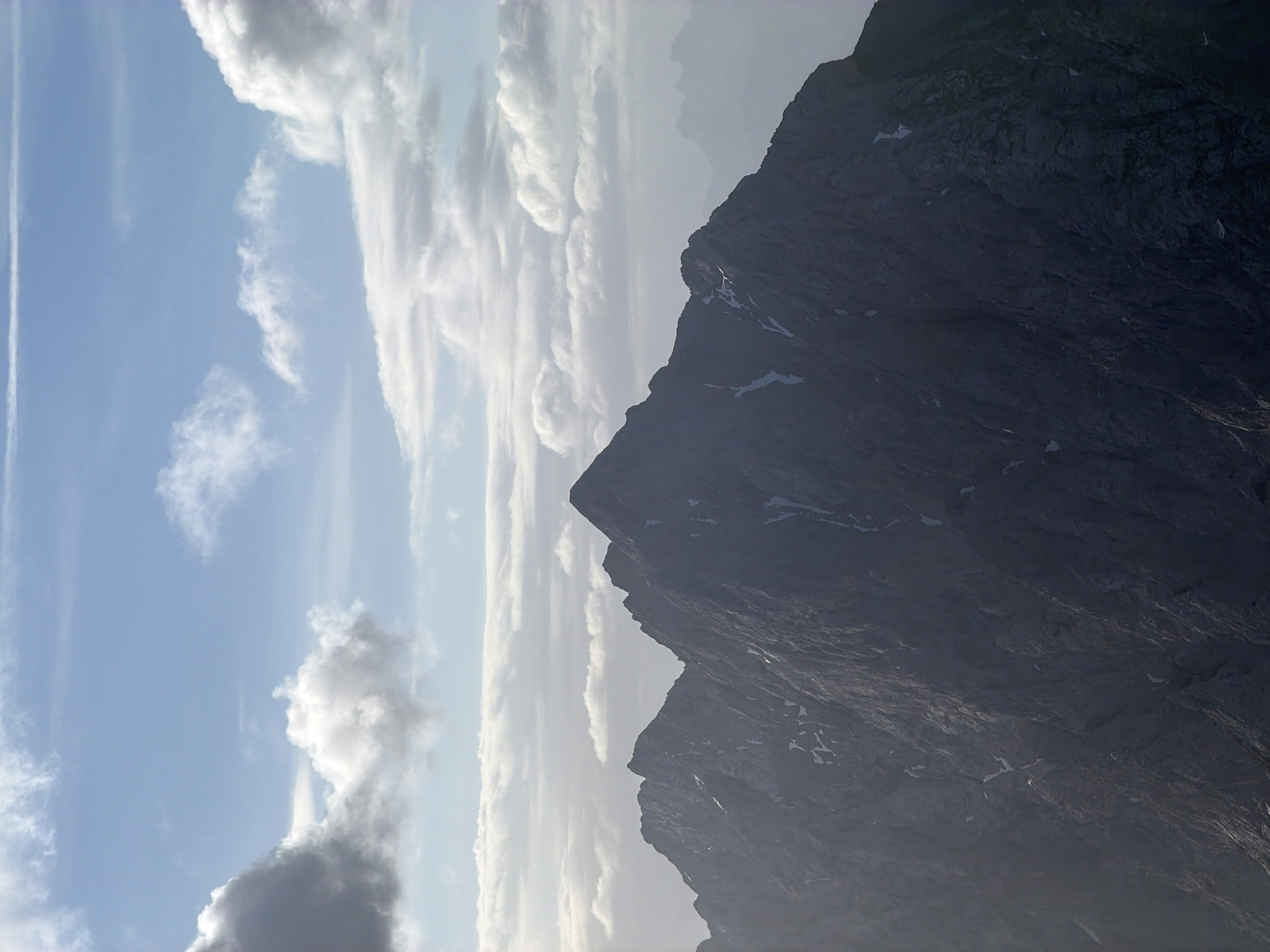 This is a landscape photograph of a mountain range under a partly cloudy sky. The mountains are rugged and appear to be made of rock, with some patches of snow or ice visible on their surfaces. The sky is a light blue with scattered white and gray clouds. In the distance, faint outlines of additional mountains can be seen through a haze. The lighting suggests it is daytime, possibly morning or afternoon. The scene appears to be a natural, remote environment.