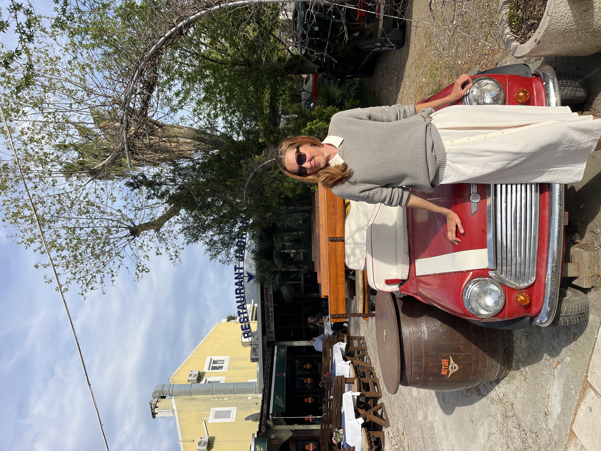 A person is leaning on a vintage, red, short-bodied car with a white stripe, parked outside the Restaurant Jadran Seaside in Budva, Montenegro. The car is mounted on a large wooden barrel with "MOTOR" inscribed on it. Tables and chairs with white tablecloths are set up outside the restaurant, and other people are visible sitting at tables. The building is yellow with visible air conditioning units. A tree canopy provides shade, and the sky is partly cloudy.