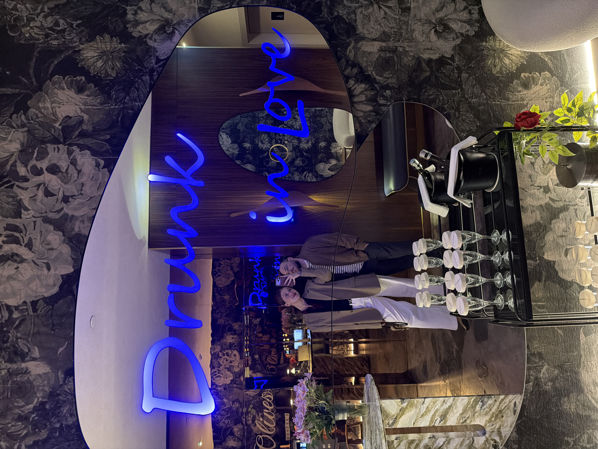A vertical indoor shot depicts a mirrored wall with blue neon writing spelling out "Drink Live Love" in a stylized font. In the mirror's reflection, two people are posing, possibly for a selfie, facing each other in front of the wall. The person on the left has fair skin, wearing a beige jacket over a dark top and white pants. The person on the right has dark hair, wearing a beige jacket over a striped shirt and dark pants. The mirror reflects the restaurant's interior, including tables, chairs, and floral arrangements. There are champagne flutes and bottles in an adjacent black rack. The wallpaper surrounding the mirror has a dark floral pattern. The location is likely Olivese Restaurant in Tbilisi, Georgia, based on the reflected signage in the mirror.