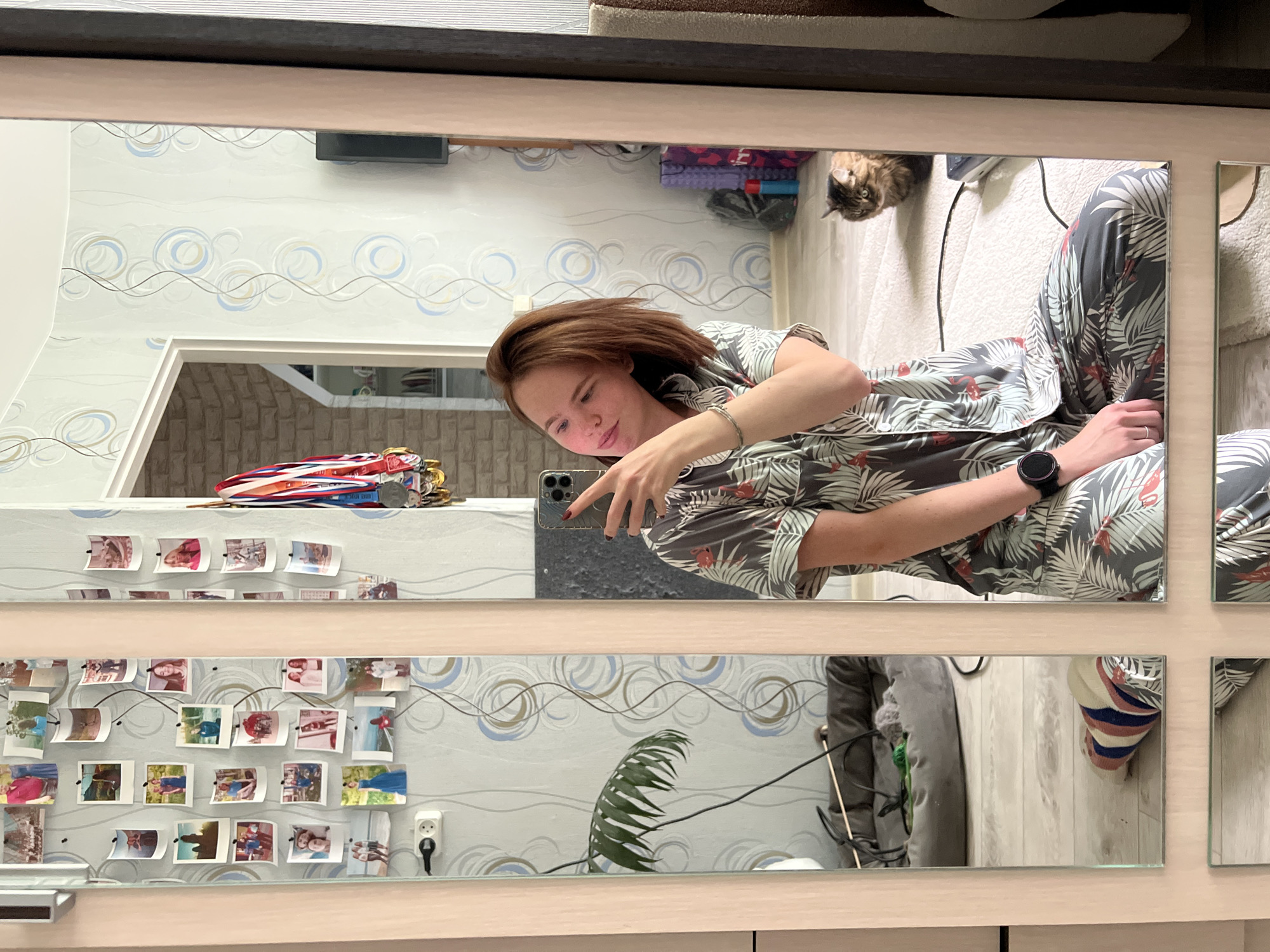 A person is taking a mirror selfie in a room. The person has short, brown hair and is wearing a grey and white patterned shirt and matching pants. They are holding a smartphone in their right hand, which is angled towards the mirror. A black smartwatch is visible on their left wrist.
The room appears to be a bedroom or living area, with patterned wallpaper featuring blue and grey swirls. In the background, a cat can be seen lying on a bed or couch. There are several framed photographs or prints displayed on the wall in the reflection. Some of the photos appear to be of people, possibly friends or family. A pile of medals is also visible on a shelf.
The lighting suggests it could be daytime, as natural light seems to be present, though there are also artificial lights in the scene. The overall mood appears casual and relaxed.
