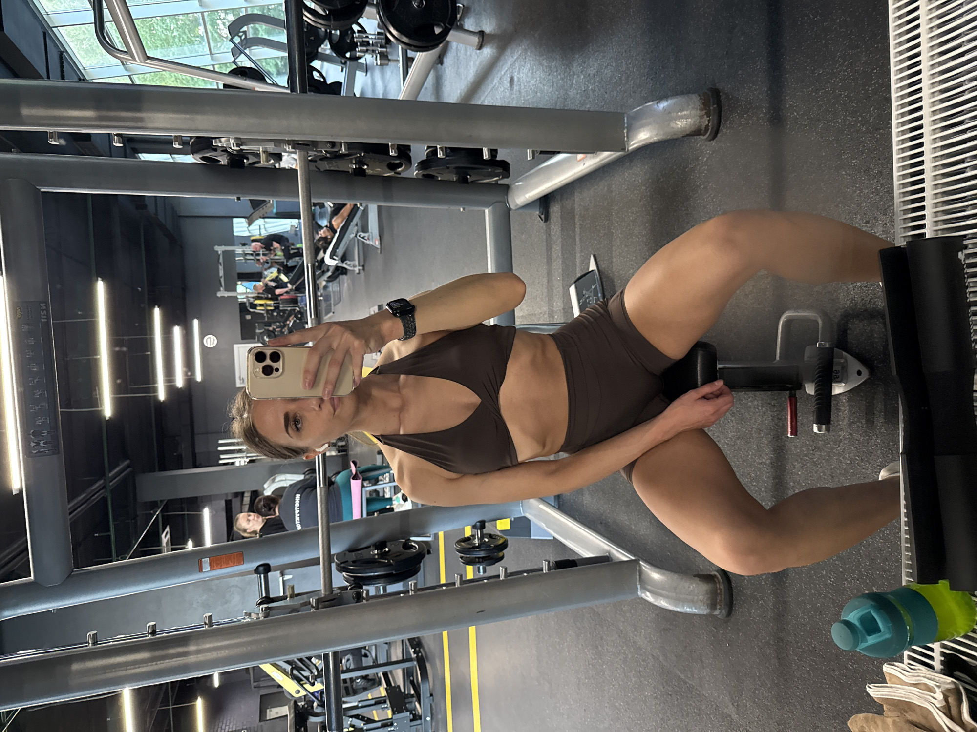 A young woman with light brown hair is seated on a black gym bench, taking a mirror selfie with a light-colored smartphone held in her right hand, partially obscuring her face. She wears a dark brown halter-neck sports bra and matching shorts, white wireless earbuds, and a dark smartwatch on her left wrist. In the foreground, a green and yellow water bottle rests on a white metal grate next to brown towels. The woman is positioned in front of a gray Smith machine with a barbell and weight plates attached to the rails. The background, largely visible through the mirror's reflection, depicts a spacious gym with dark gray rubber flooring, yellow markings, and overhead rectangular light fixtures. Various gym equipment, including benches, weight racks, and other exercise machines, are present. Several other individuals are visible in the background, some engaged in exercise or conversation. Large windows on the right side of the reflection reveal green foliage outside, indicating a daytime setting. This scene is located within a gym in Saint Petersburg, Russia.