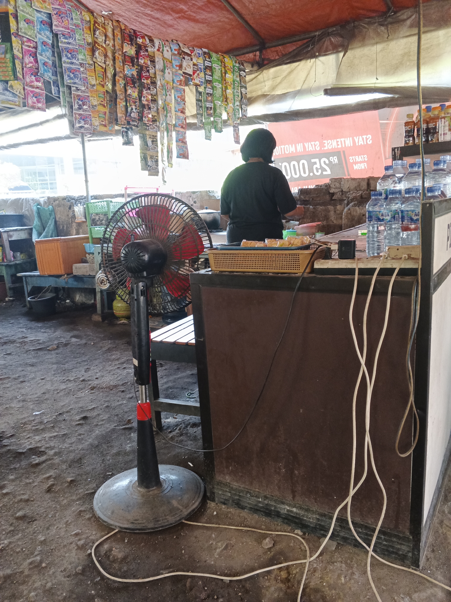 In Tambak Kemerakan, Indonesia, an outdoor market stall displays an array of bagged goods hanging from the red awning. A person, seen from behind wearing a black t-shirt, is attending to the stall. In the foreground, a black and red oscillating fan stands on a dusty ground. The stall counter is laden with several bottles of Aqua mineral water and a tray of food items. Electrical cords are visible, extending from an outlet strip on the counter and across the floor. The background reveals a rough stone wall and other market stalls or structures.