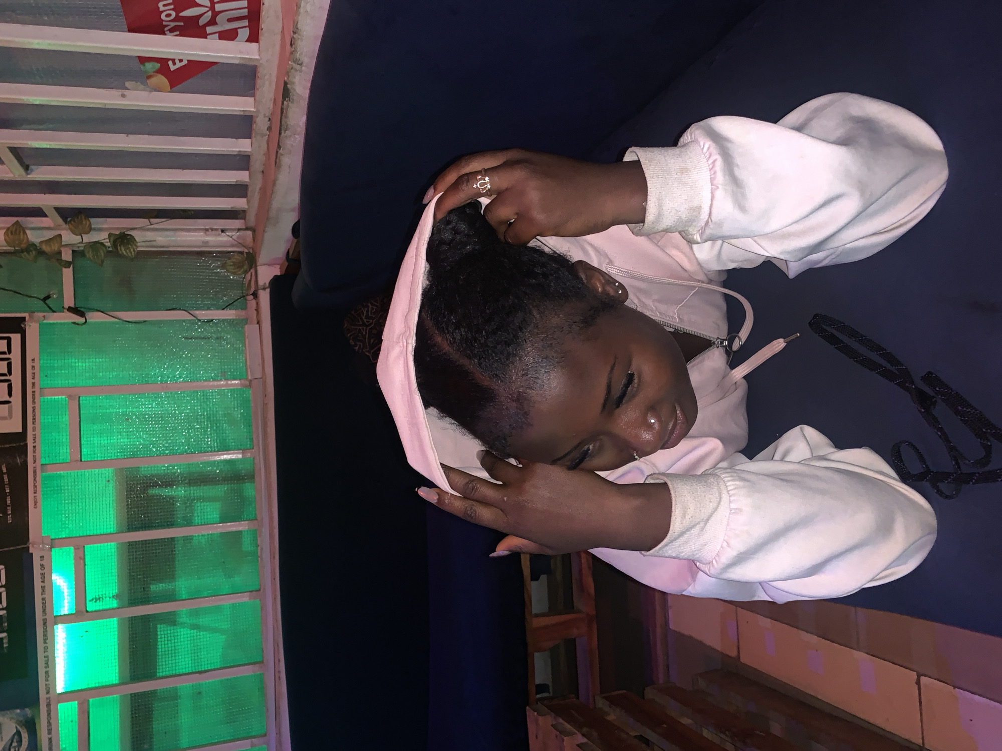 A young woman is depicted resting or sleeping on a dark blue surface, likely a sofa or bench, in what appears to be an indoor or sheltered outdoor setting. She has dark skin, with her braided hair pulled back, and is wearing a light pink hooded sweatshirt with white ribbed cuffs. Her eyes are closed, and her head is tilted to the side, suggesting peaceful slumber. Notable accessories include a ring on her right ring finger and small piercings on her nose and ear.
To the left of the woman, a distinctive structure with white horizontal bars and translucent green panels forms a partition or window, illuminated by a green light source. Artificial green leafy vines are draped along the top of this structure. Above this, a red sign with white text partially reads "Everyon Chi," possibly "Everyone's Chill." Below this, a white label warns, "ENJOY RESPONSIBLY NOT FOR SALE TO PERSONS UNDER THE AGE OF 18" and "DRINK RESPONSIBLE NOT FOR SALE ONS UNDER THE AGE OF 18," indicating an establishment that serves alcohol. A black strap or cord is visible near her right arm. In the foreground, a wooden structure, possibly made of pallets, can be seen. The ambient lighting suggests an evening or nighttime scene. The location is consistent with a bar, lounge, or similar venue in Barakin Akawo, Nigeria.