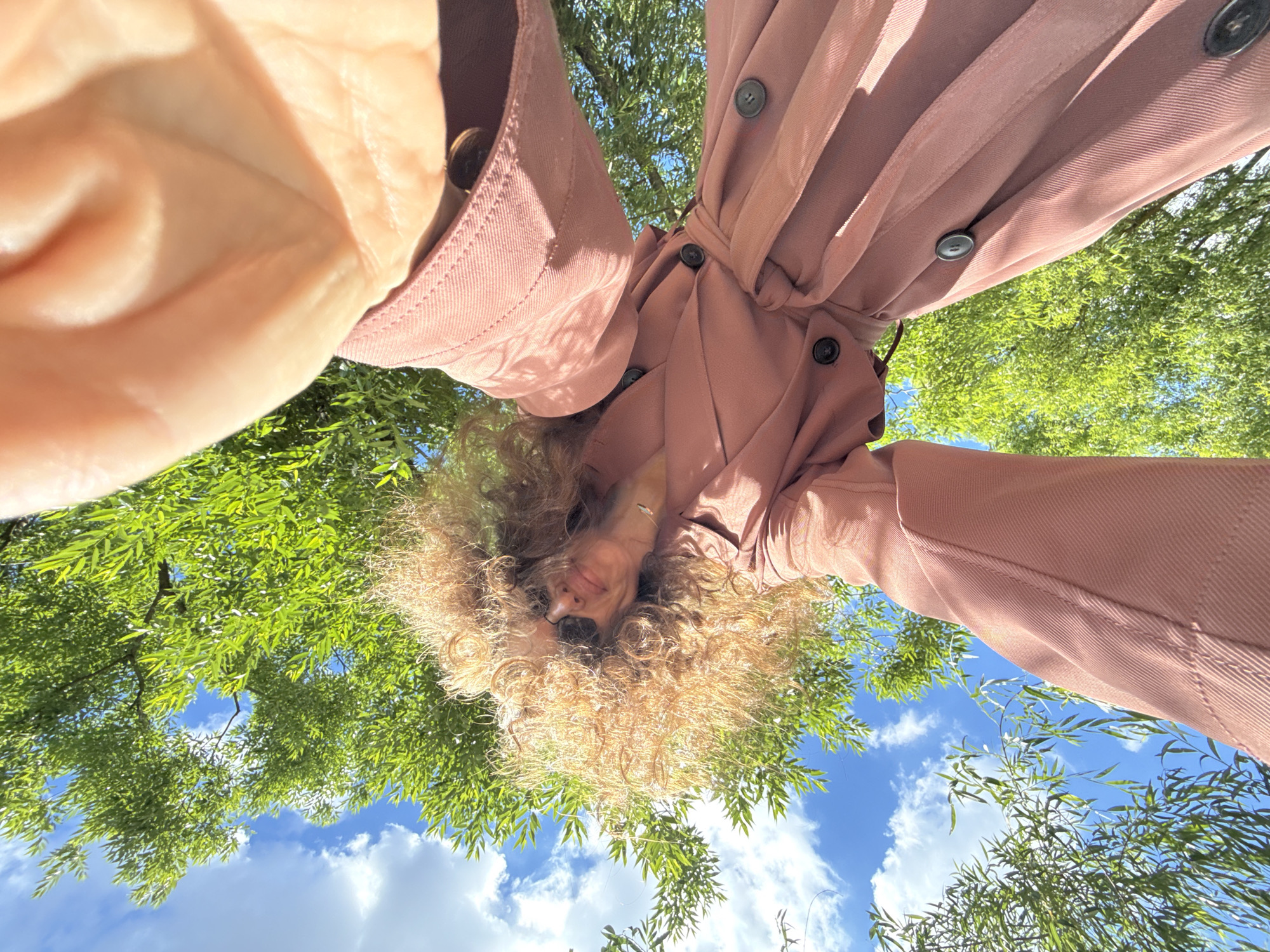This is a low-angle, upside-down selfie taken outdoors. The subject is a person with curly, blonde hair wearing a light pink button-up shirt. They are looking directly at the camera with a slight smile. Their face is framed by their hair and the pink fabric of their shirt. In the background, lush green leaves of trees are visible against a bright blue sky with scattered white clouds. The lighting suggests it is daytime, likely sunny. The perspective makes it appear as though the person is looking up from beneath the trees. There is no visible text in the image. The overall scene conveys a sense of warmth, nature, and casual outdoor activity.