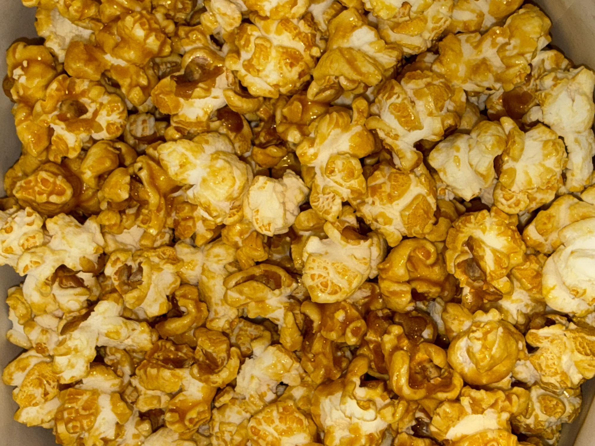 The image shows a close-up, top-down view of a large quantity of caramel popcorn. The popcorn pieces are clustered together, with the caramel coating creating a glossy, golden-brown sheen over the white, puffy kernels. The texture appears slightly sticky and crunchy. There are no people, actions, or environmental details visible, as the image is solely focused on the food item. The scene is a generic presentation of popcorn and does not provide any information about its location in Dubai, United Arab Emirates, or any other specific place.