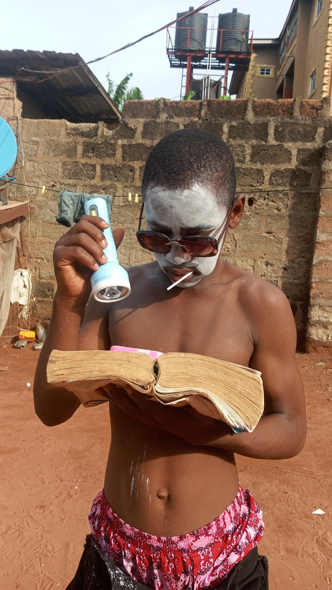 A shirtless boy with sunglasses and white face paint stands outside, holding a flashlight in his left hand and an open book in his right. He has a lollipop in his mouth. The background includes brick buildings, water tanks, and red dirt ground. The location is Unknown, Unknown.