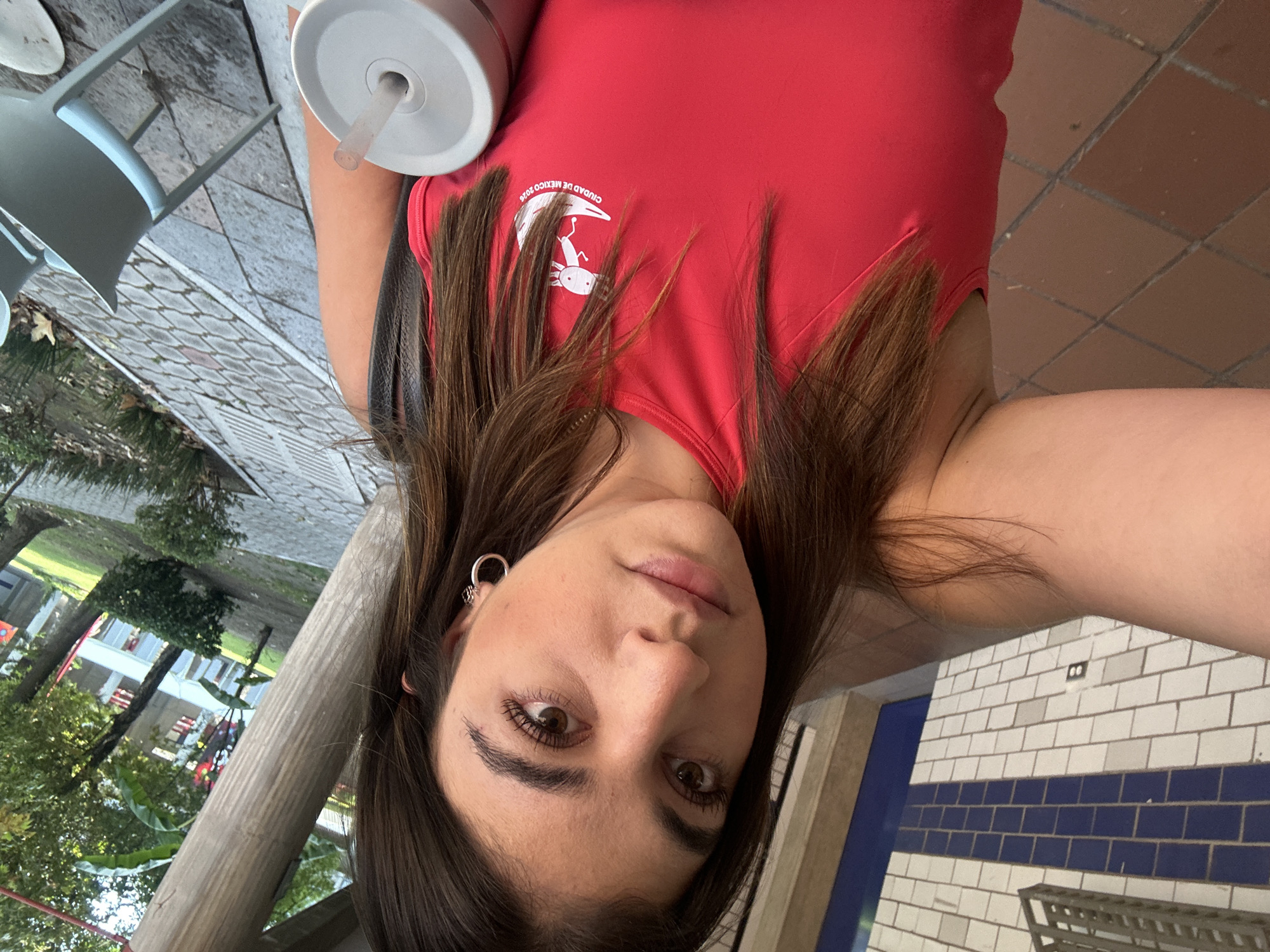 A person is shown in a close-up, angled selfie. The subject has brown hair, light skin, and brown eyes. They wear a red top with a white logo depicting an axolotl and the words "CIUDAD DE MEXICO 2026". An earring is visible in their left ear. They appear to be holding a cup with a gray lid and straw.
The background includes a mix of indoor and outdoor elements. Outdoor elements visible are trees and building. A brick tiled floor and white brick wall with a blue stripe are also visible.
I cannot confirm the exact location in San Bernardino Tlaxcalancingo, Mexico.