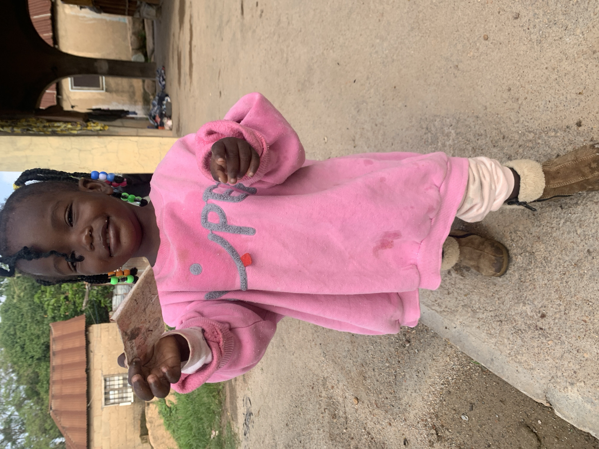 A young Black girl with braided hair decorated with colorful beads smiles at the camera while holding a piece of debris in her outstretched right hand. She is wearing a pink sweatshirt with gray lettering, light pink leggings, and brown fur-lined boots. The girl is standing on a dirt and concrete path in an outdoor, rural setting, likely in Jos, Nigeria. In the background, there are simple buildings with corrugated metal roofs and arched doorways, suggesting a modest village or community. The ground is a mix of dirt, gravel, and some scattered debris. The overall scene depicts a child in a developing environment.